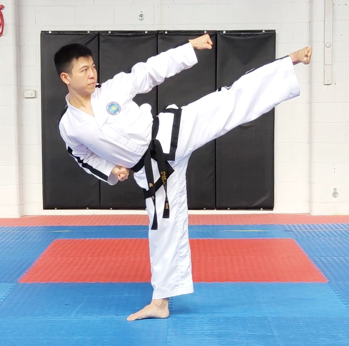 Martial artist in white gi and black belt holding a high side kick on a blue-and-red mat