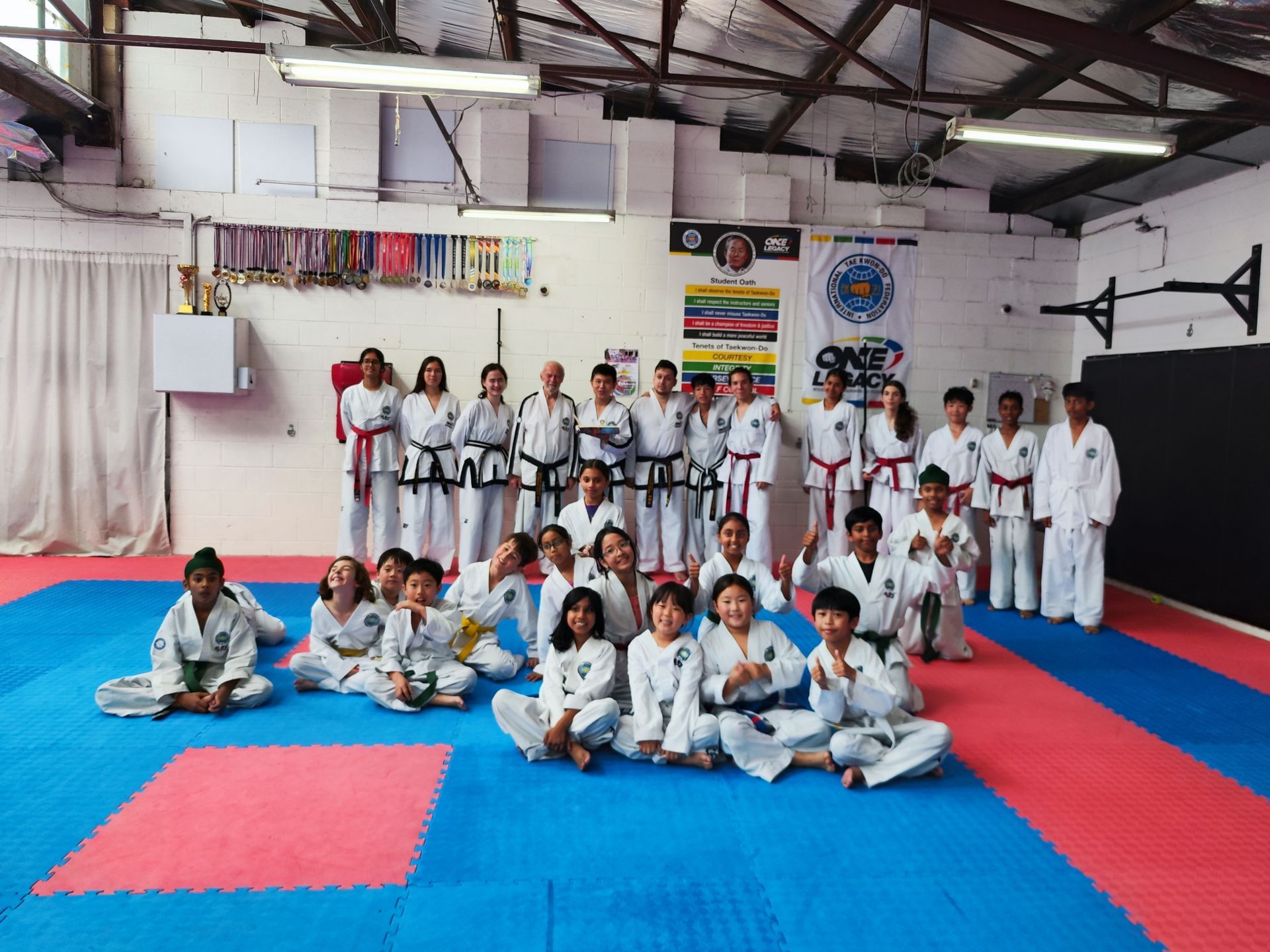 Martial arts class posing on blue and red mats in a dojo with white uniforms and belts displayed on the wall