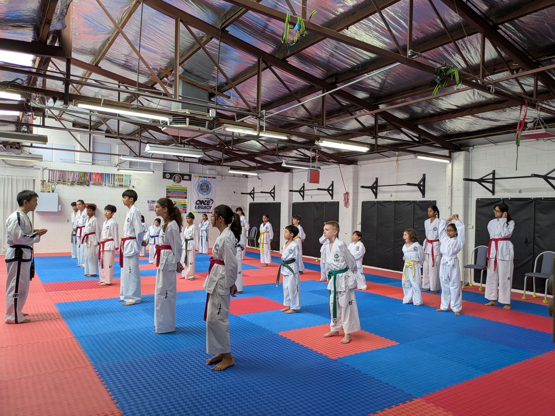 Martial arts class lined up on blue mats in a training gym, instructor at left speaking to students.