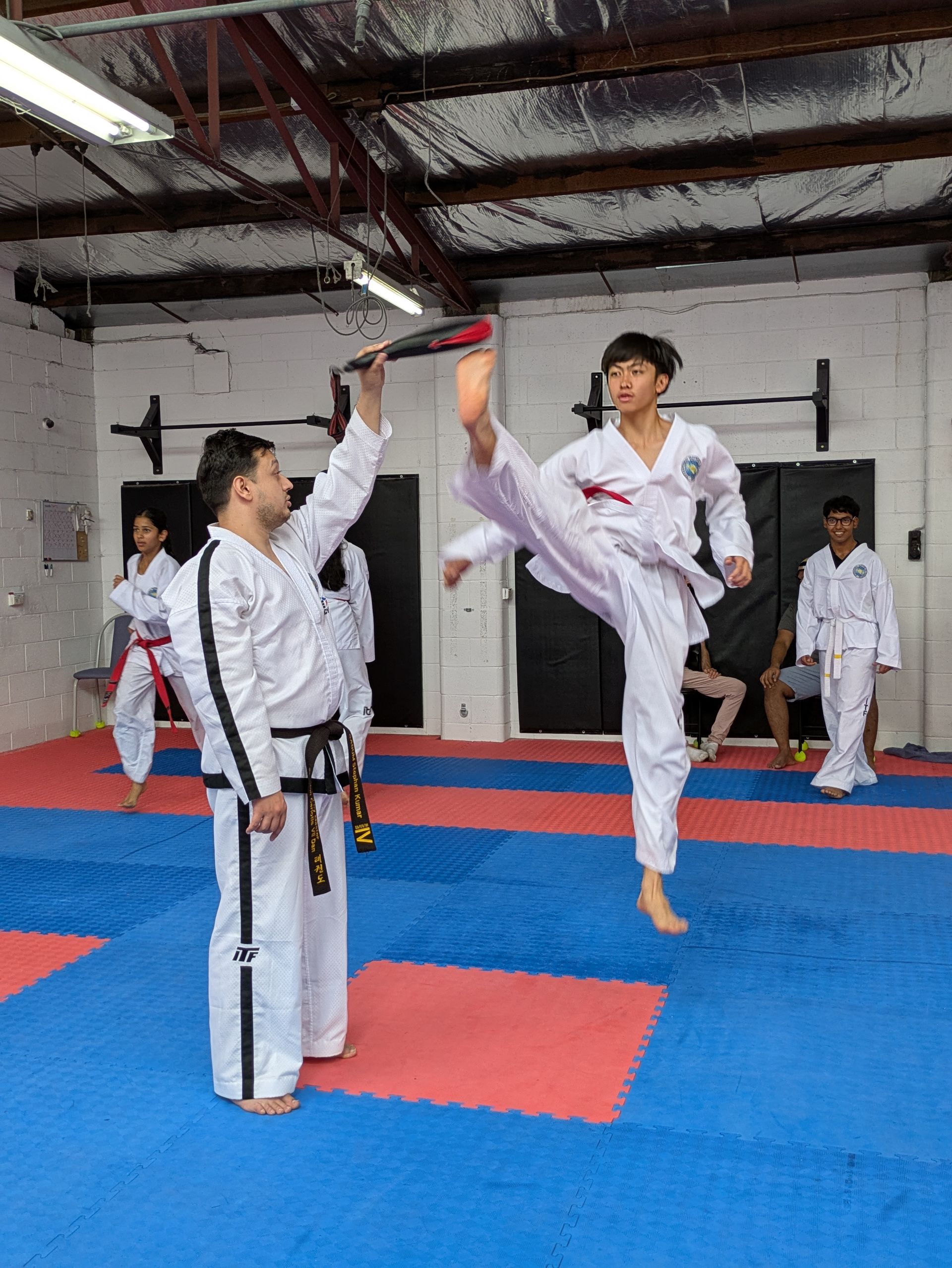Martial arts students in white uniforms practicing a high kick in a gym with blue and red mats