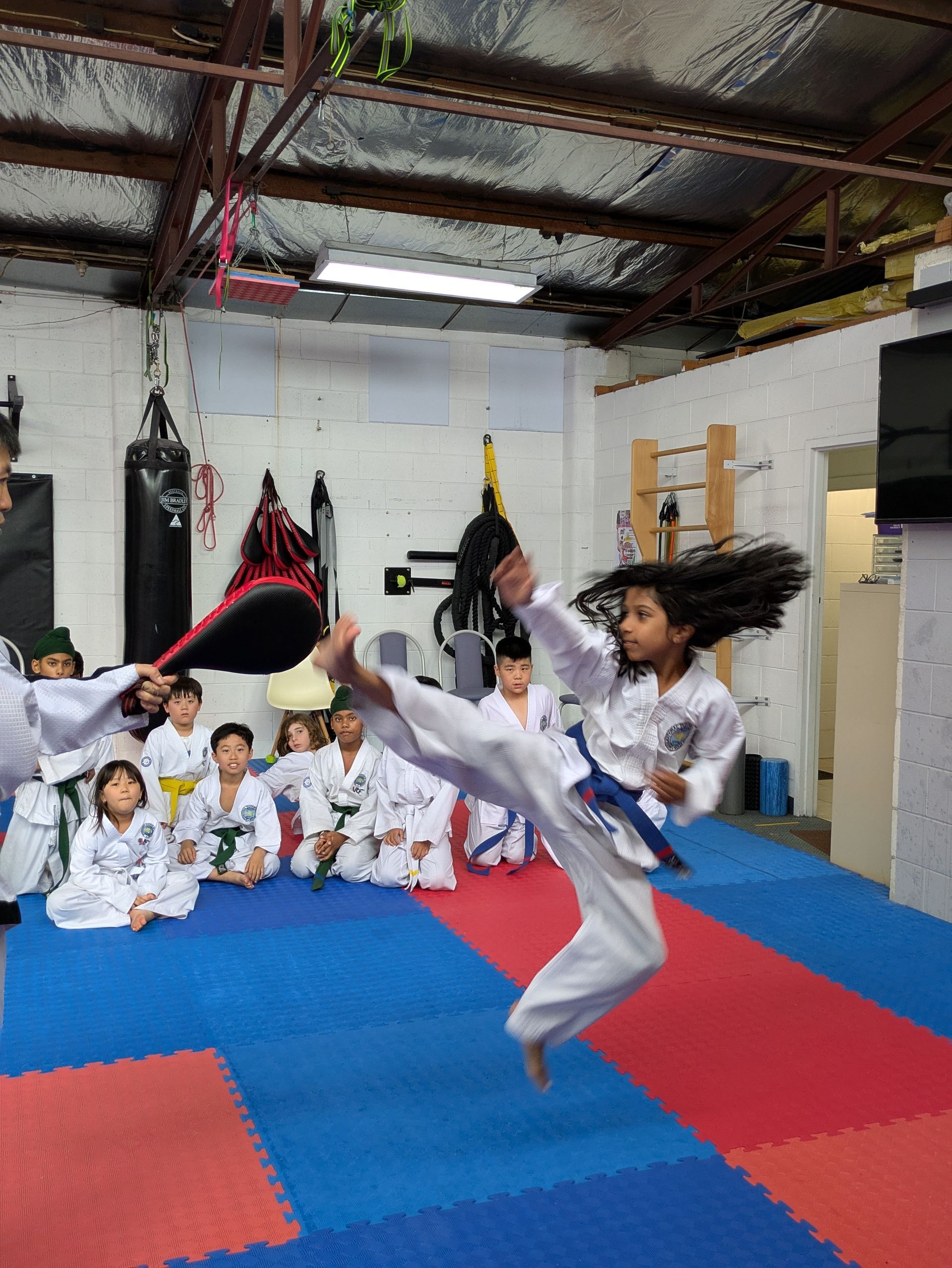 Martial arts class indoors, a student kicks high on blue mats while others watch in white uniforms