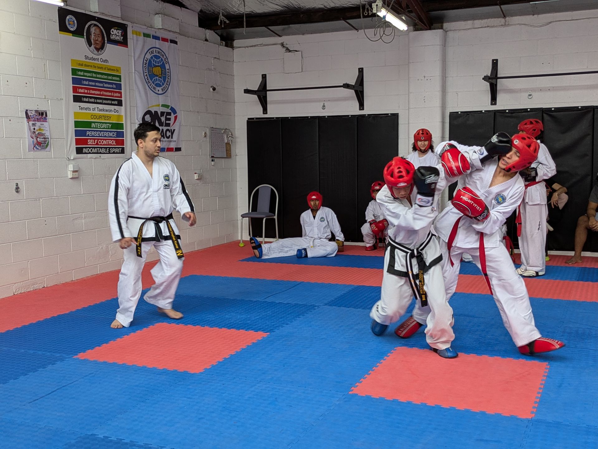 Karate class sparring on blue mats, with students in red helmets and a black belt instructor watching.