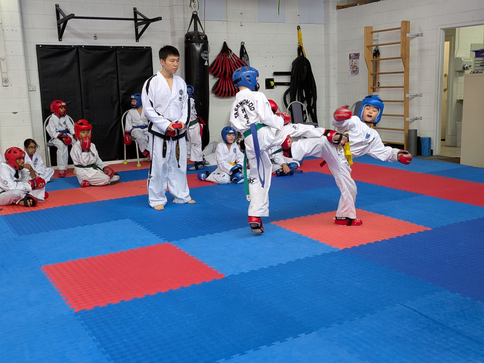 Children practicing martial arts kicks on red and blue mats in a dojo, with an instructor watching.