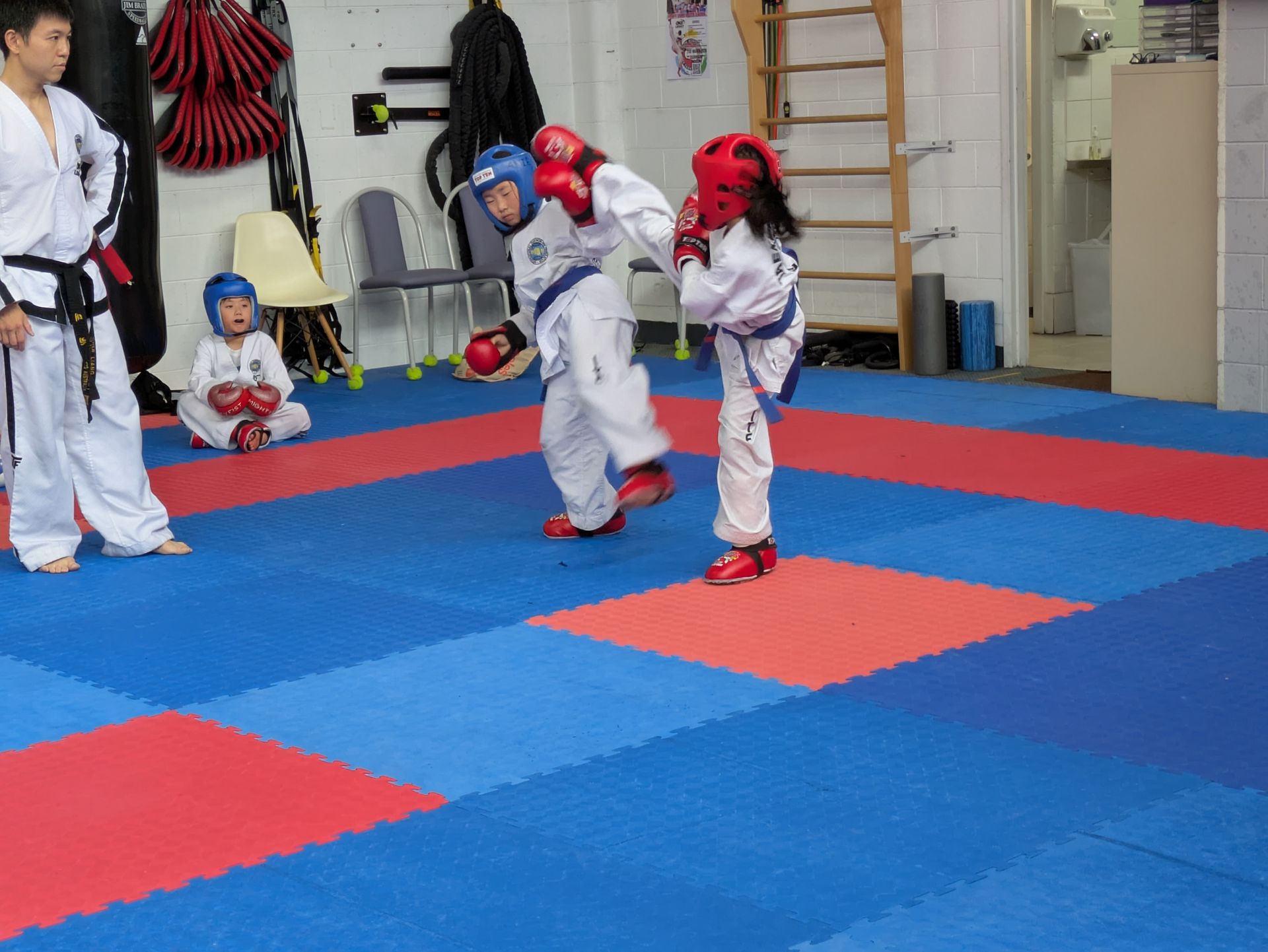 Children sparring in protective gear on blue and red mats in a martial arts gym