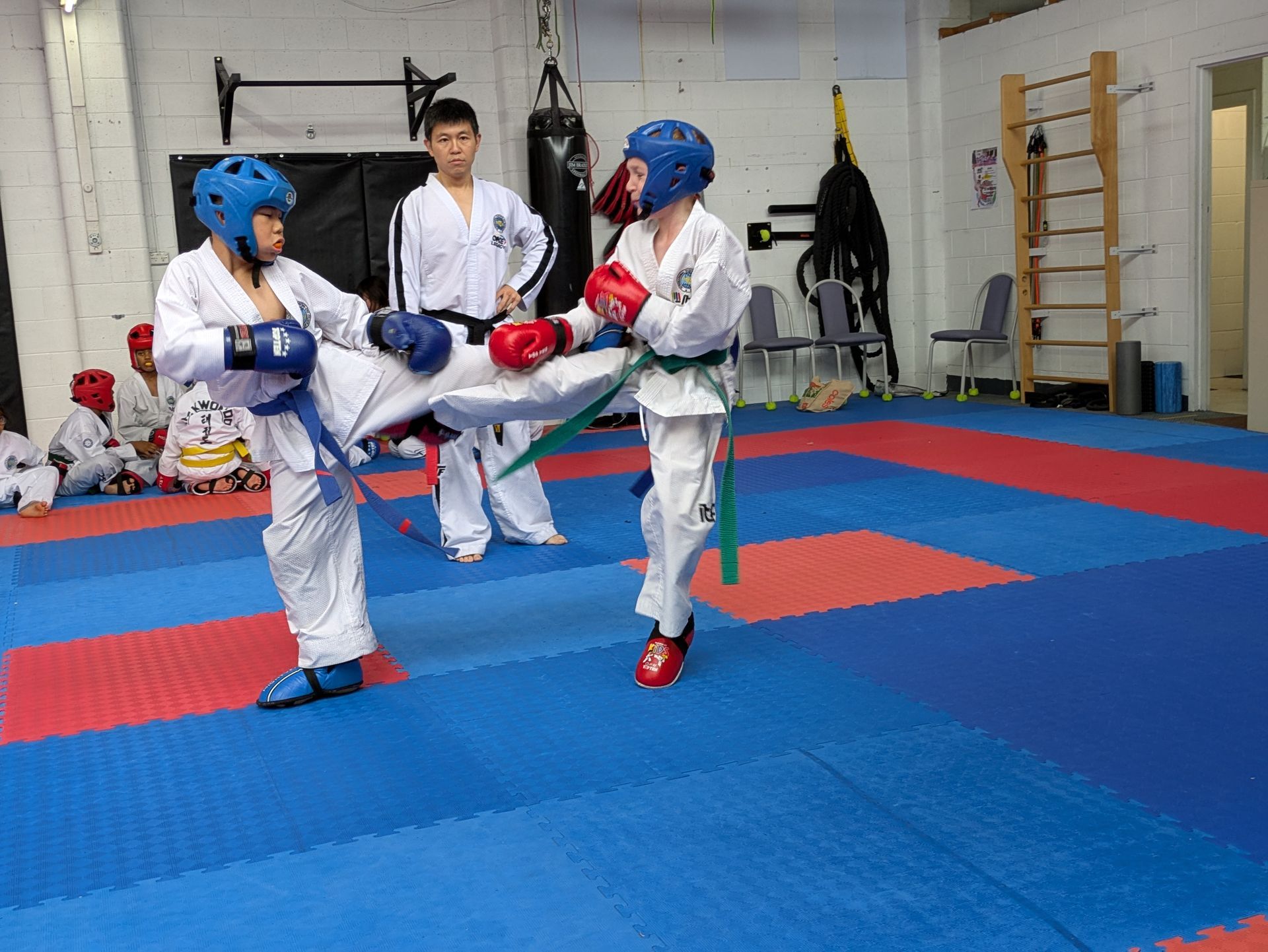Two children sparring in a martial arts gym, wearing blue helmets and gloves on red-and-blue mats.