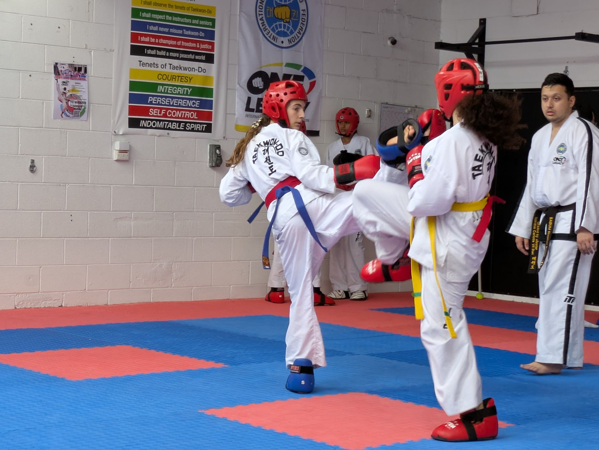 Two taekwondo sparring partners in red headgear kick in a training studio while an instructor watches.