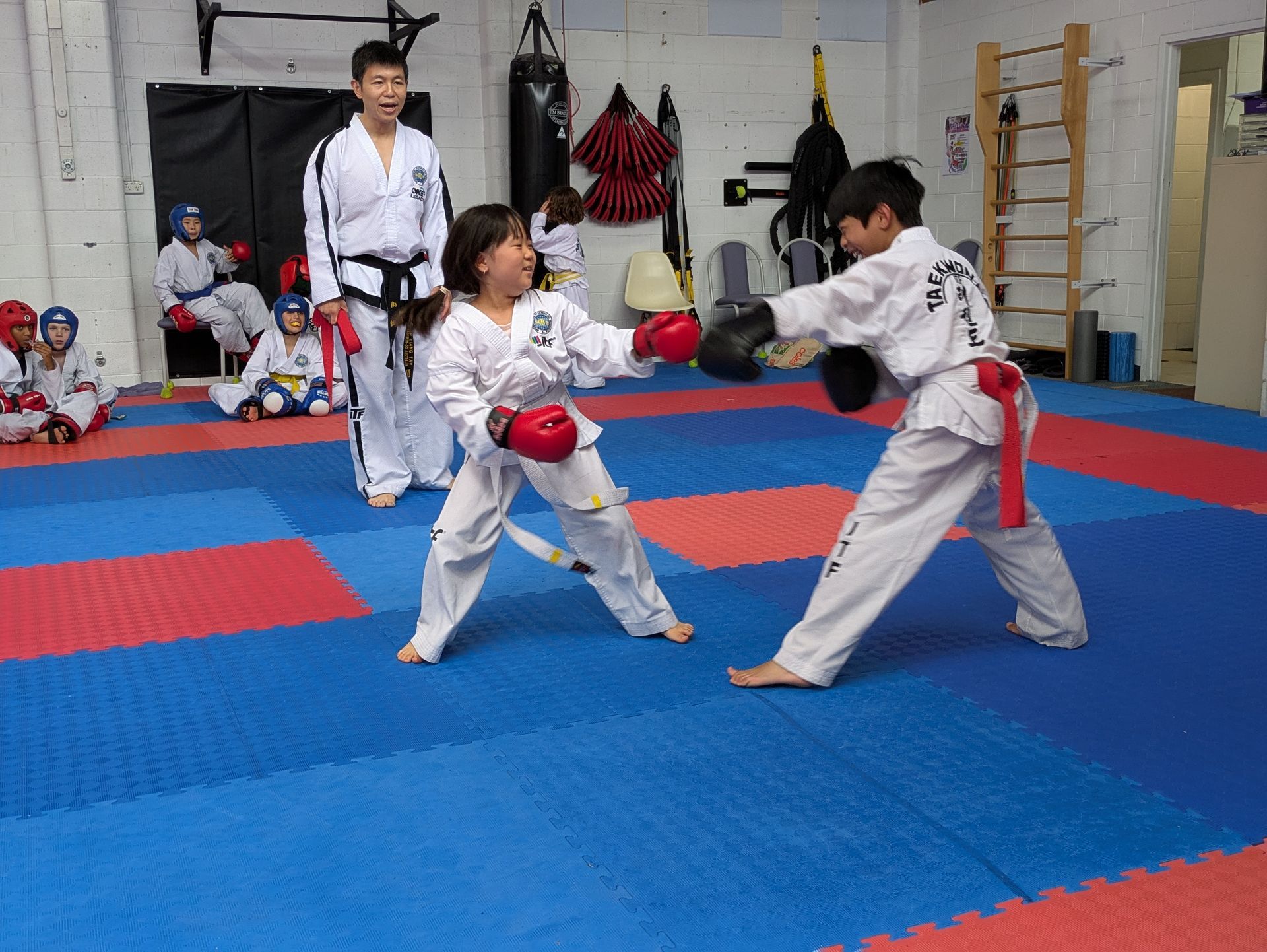 Two martial arts students spar in a dojo, with an instructor watching nearby.