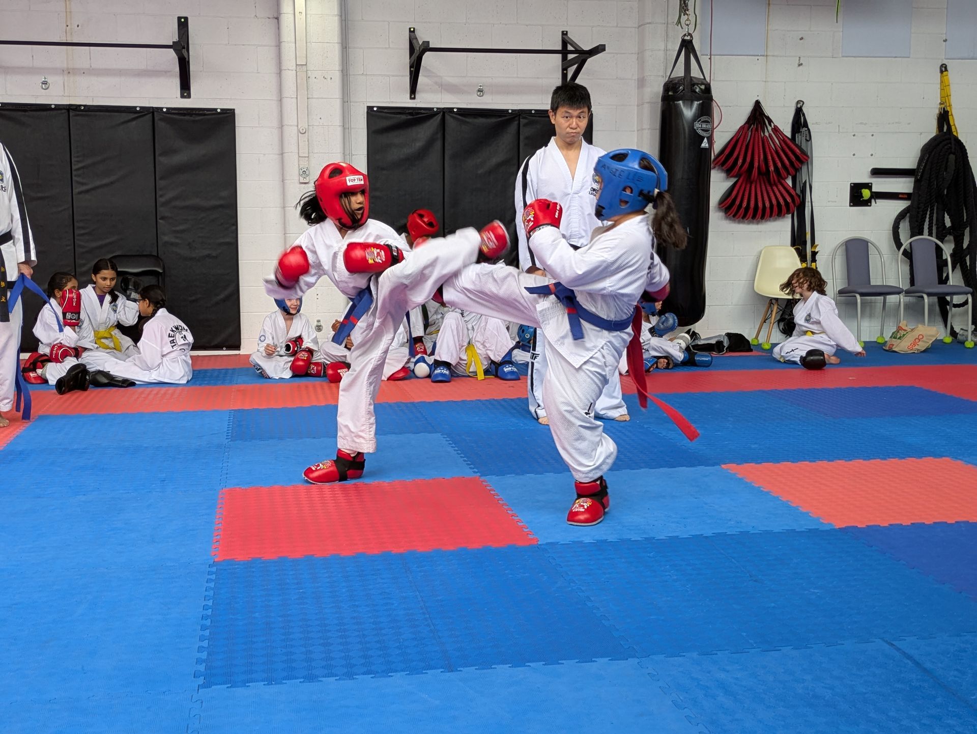 Two children sparring in taekwondo gear on a blue mat, with a coach watching nearby.