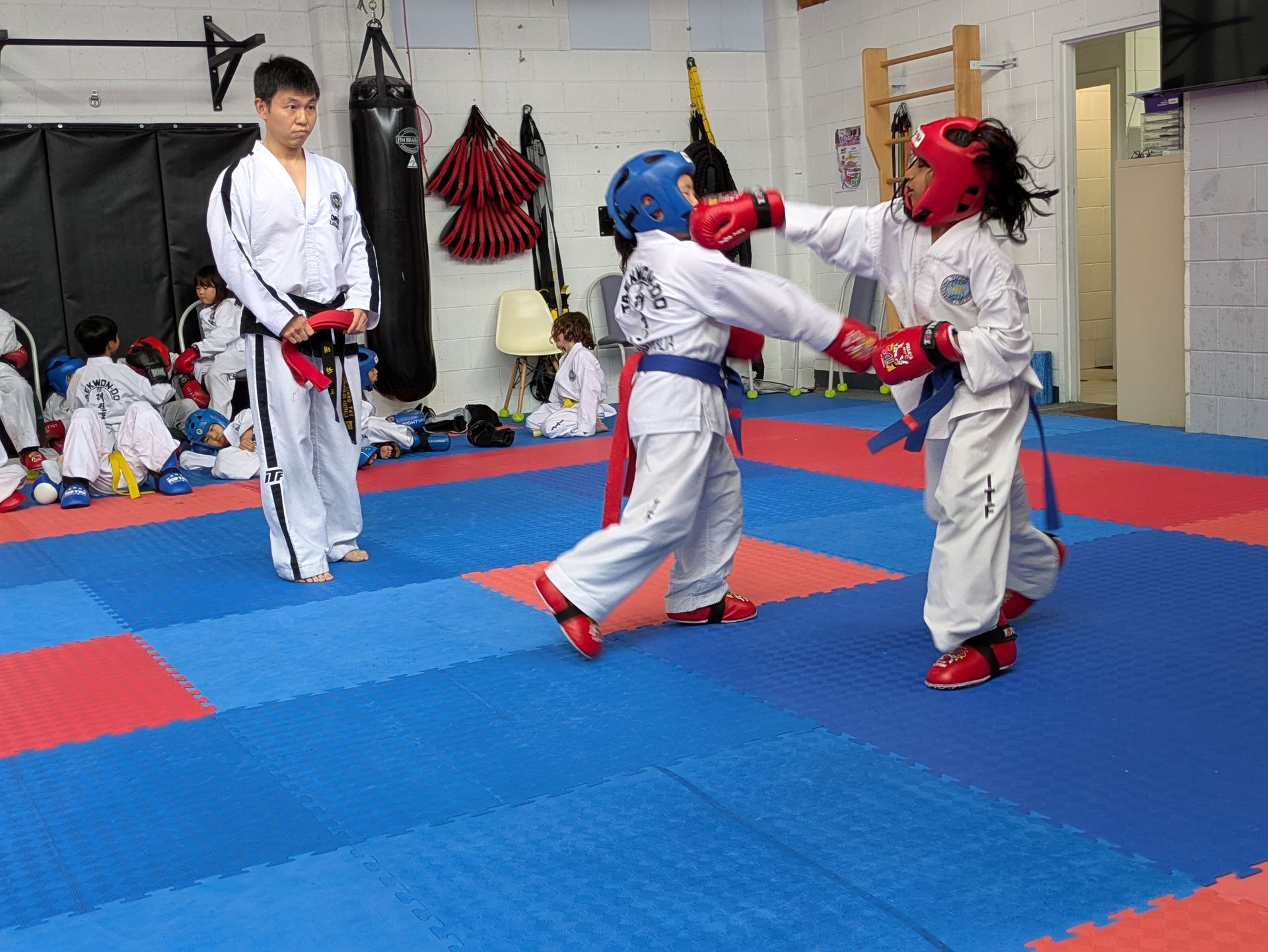 Children practicing martial arts sparring on blue mats in a gym, wearing white uniforms and red/blue protective gear