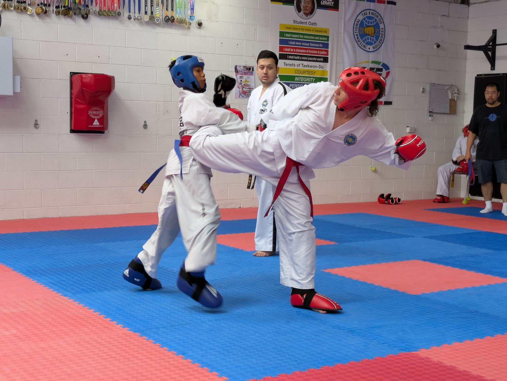 Two taekwondo sparring athletes in white uniforms and red-blue protective gear on a mat, with a referee behind them.