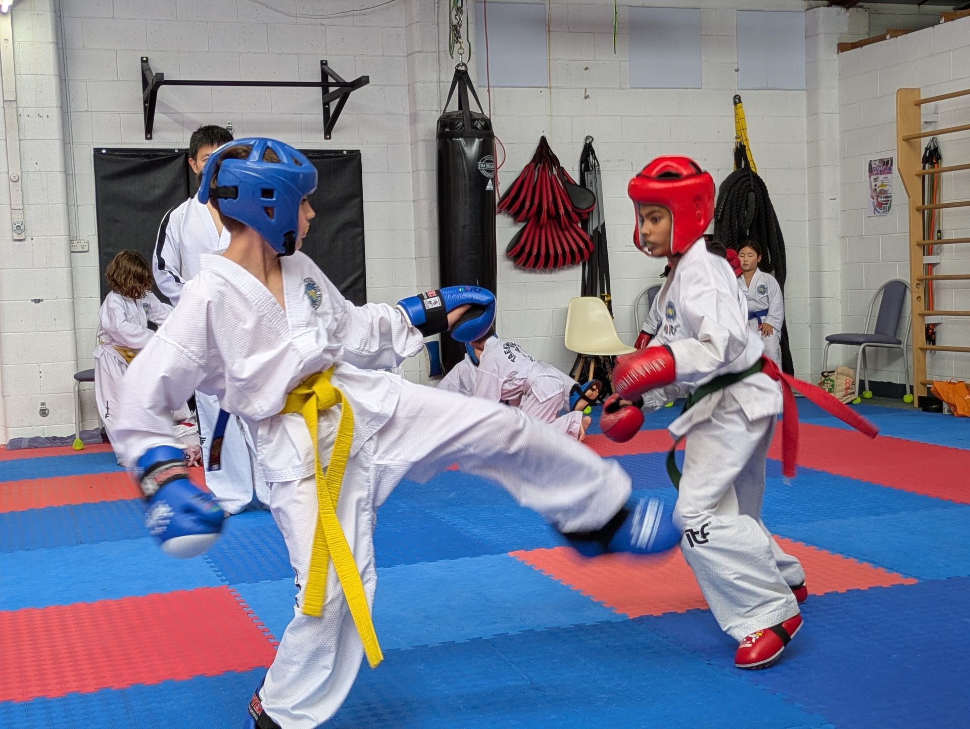 Two martial artists sparring in protective gear on blue and red mats in a gym