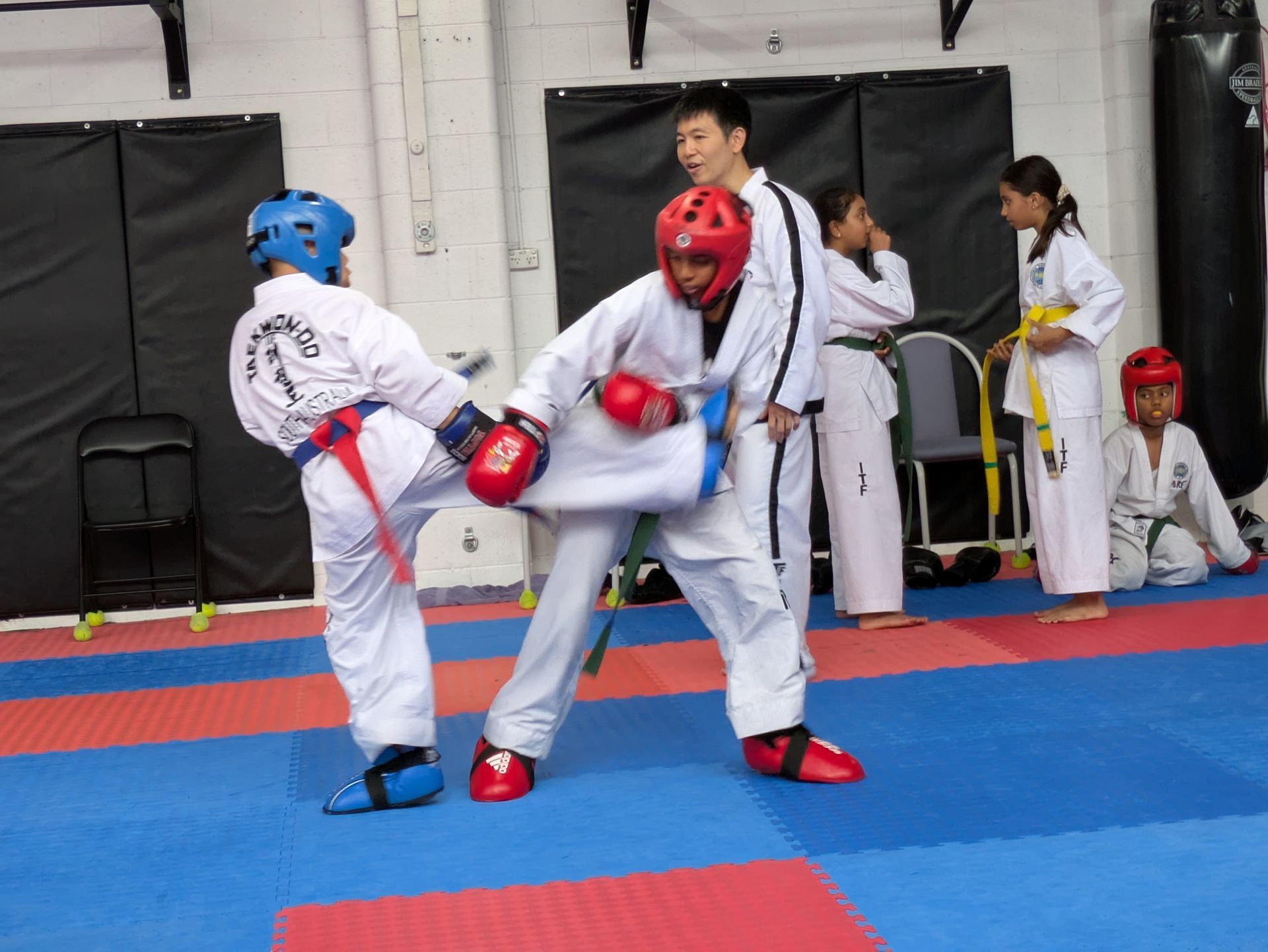 Two taekwondo sparring competitors in blue and red gear kick and block on a mat in a gym