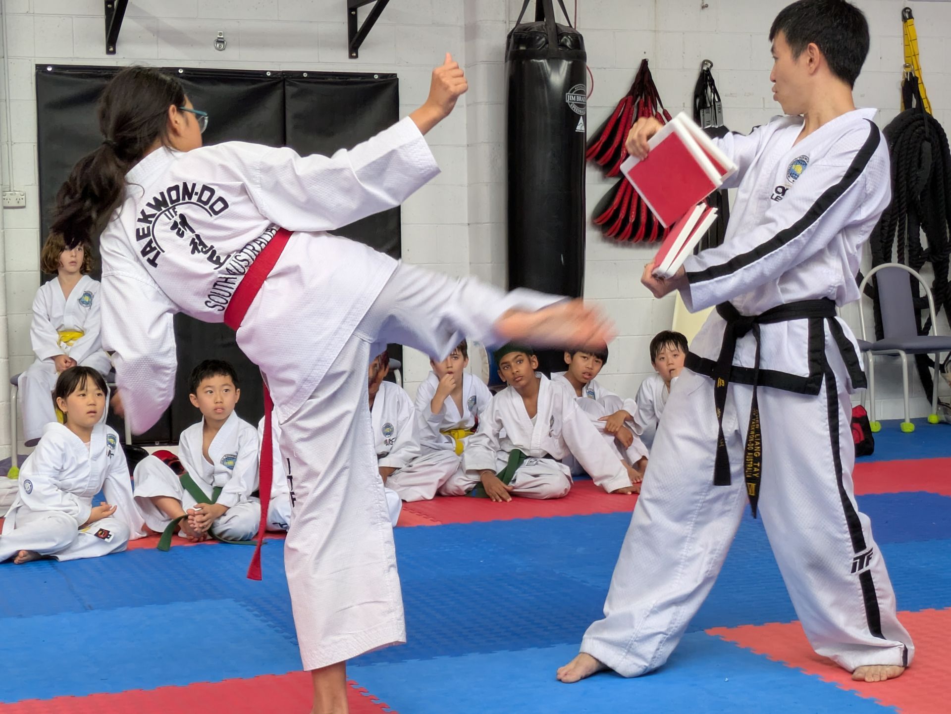 Taekwondo class: student kicks a pad held by instructor while others watch on a mat