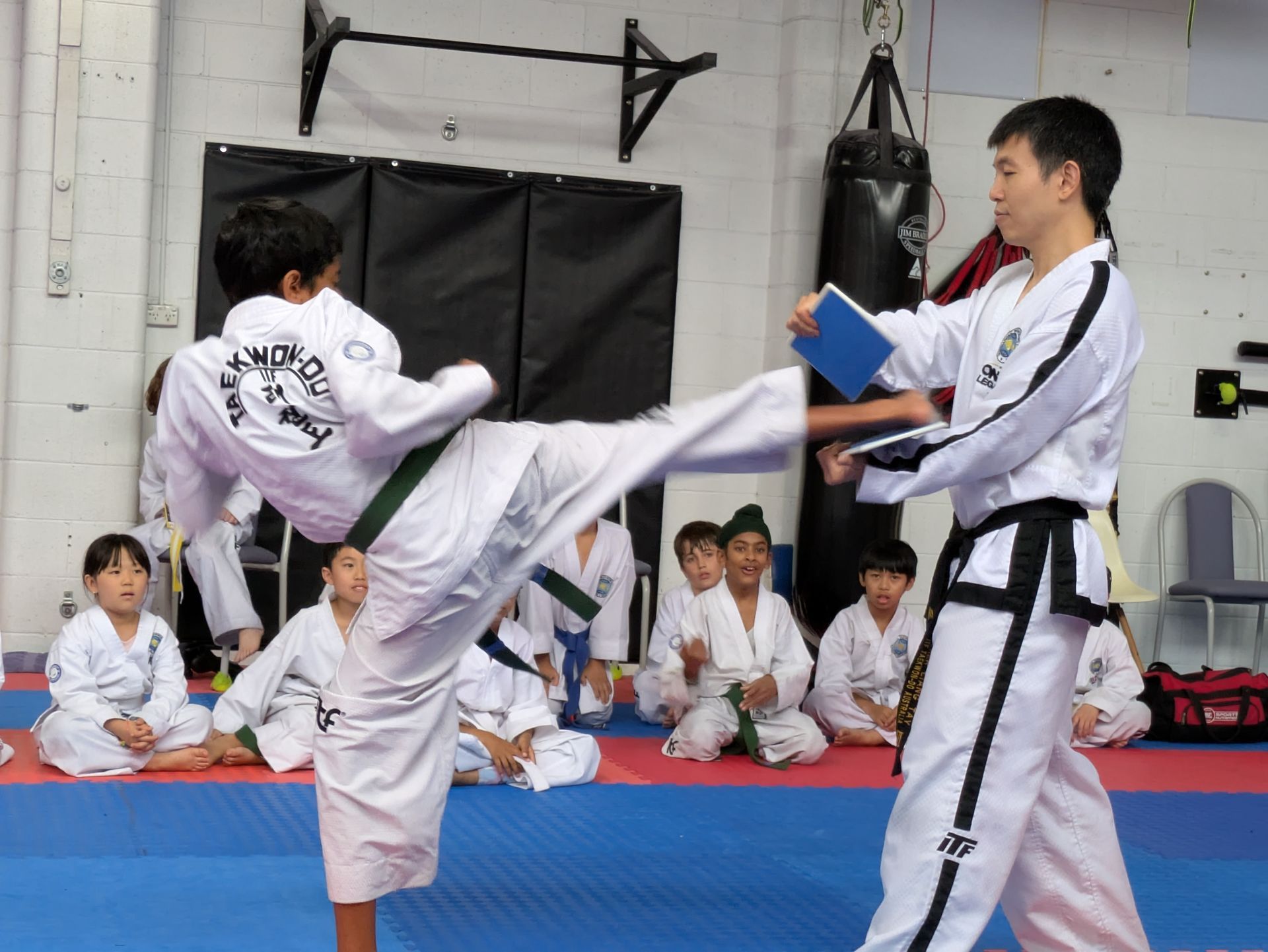 Martial arts class with a student kicking a board held by an instructor on a blue mat