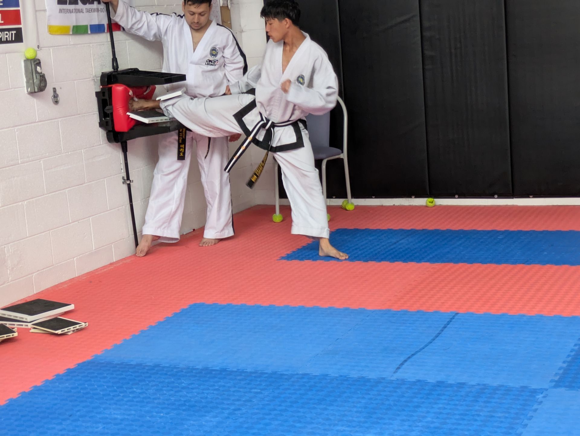 Two martial artists in white gis practicing kicks on red and blue mats in a dojo