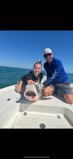 Two people kneel on a boat deck with a large fish against a bright blue ocean and sky.