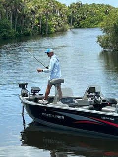 Man fishing from a black Crestliner boat on a calm, tree-lined river