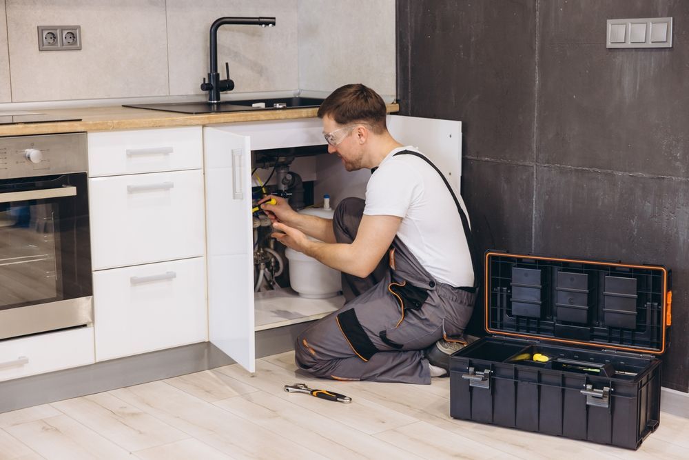 A technician in gray work clothes kneels on a kitchen floor, repairing plumbing underneath a white sink cabinet.