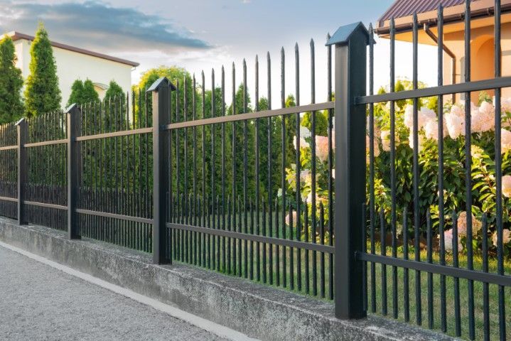 Black metal fence with vertical bars in front of a house, set on a concrete foundation, with greenery in the background.