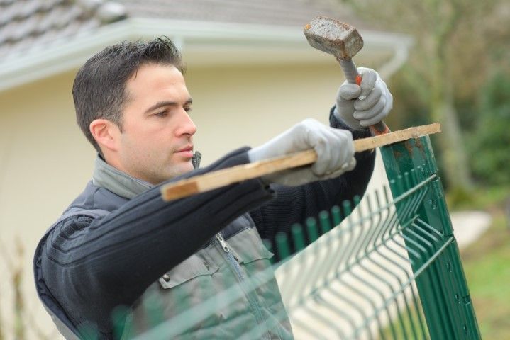 Man in gloves hammering a wooden plank on green fence in outdoor setting.