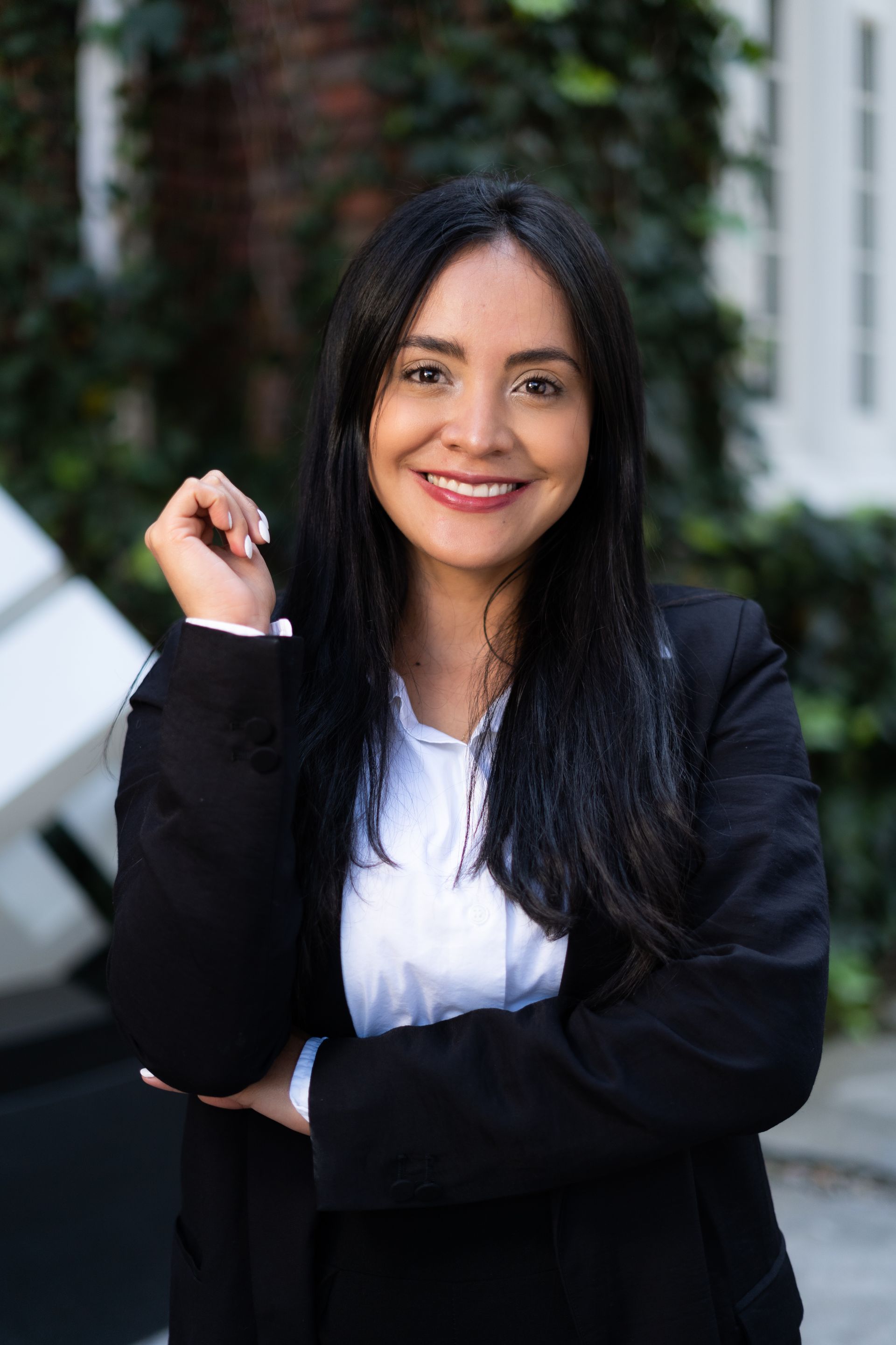 Smiling professional with long dark hair, wearing a white shirt and black blazer, posed outdoors near green foliage.