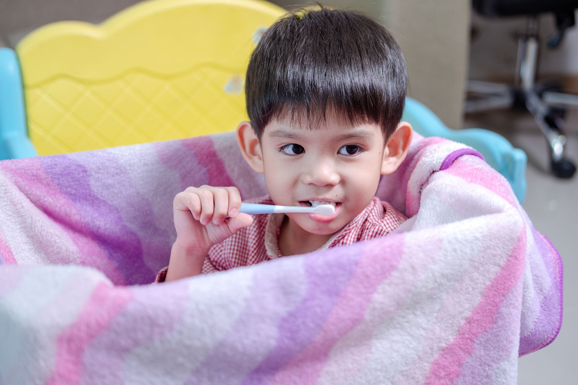 A young boy is brushing his teeth while sitting in a chair covered in a blanket.