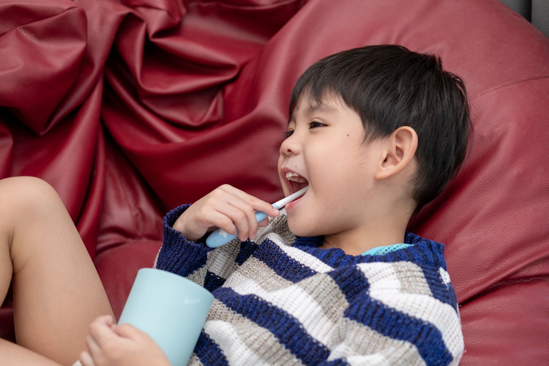A young boy is brushing his teeth while laying on a bean bag chair.
