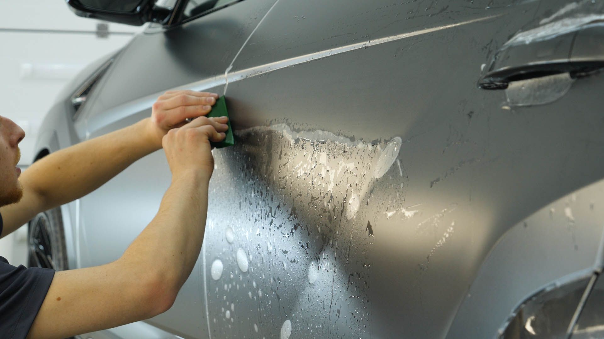 Person applying film to a gray car door with water droplets during a detailing or wrap installation.