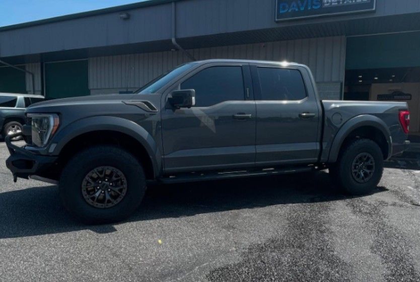 Gray Ford Raptor pickup truck parked on asphalt in front of a building with dark tinted windows.