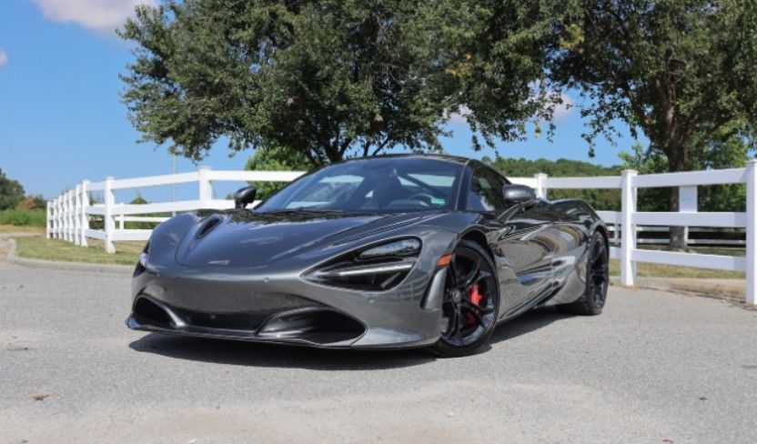 Gray McLaren sports car parked in front of a white fence and trees on a sunny day.