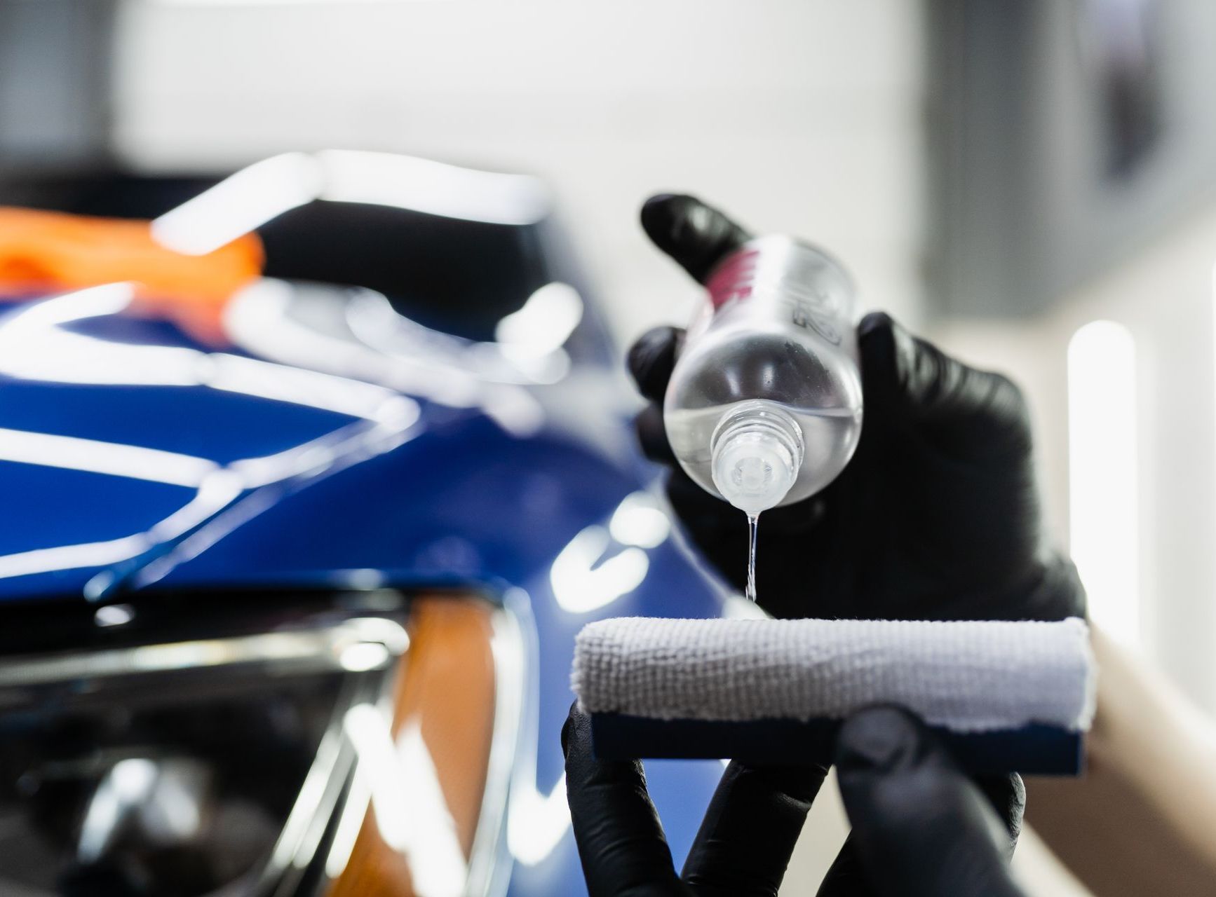 Car detailing spray bottle in front of a blue car, with a gloved hand holding a microfiber cloth