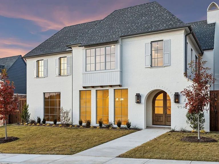 White two-story house with arched front door, large windows, gray roof, and small trees on a grassy lawn.