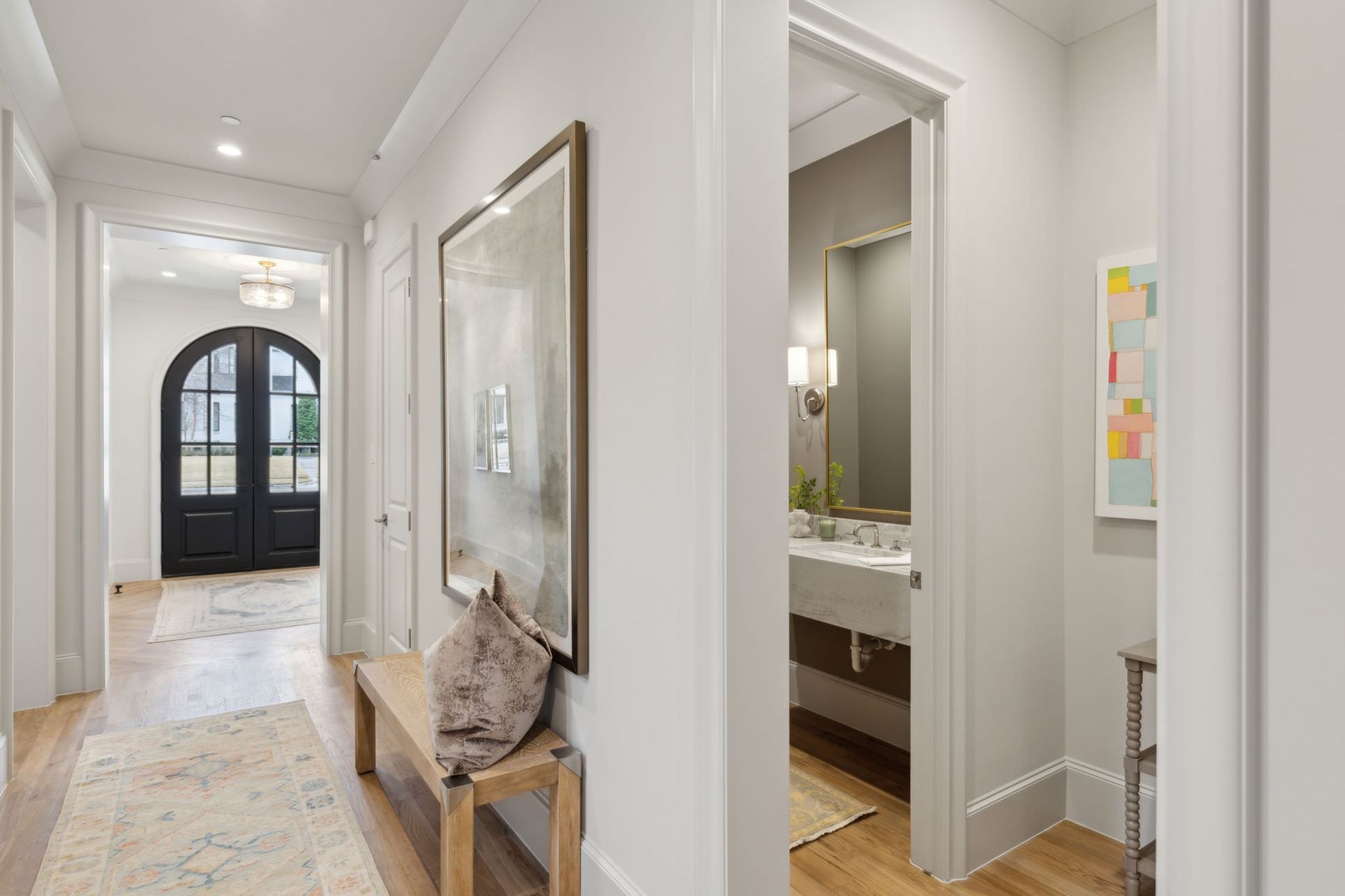 Hallway with light wood floors, a bench, artwork, and a doorway leading to a powder room.