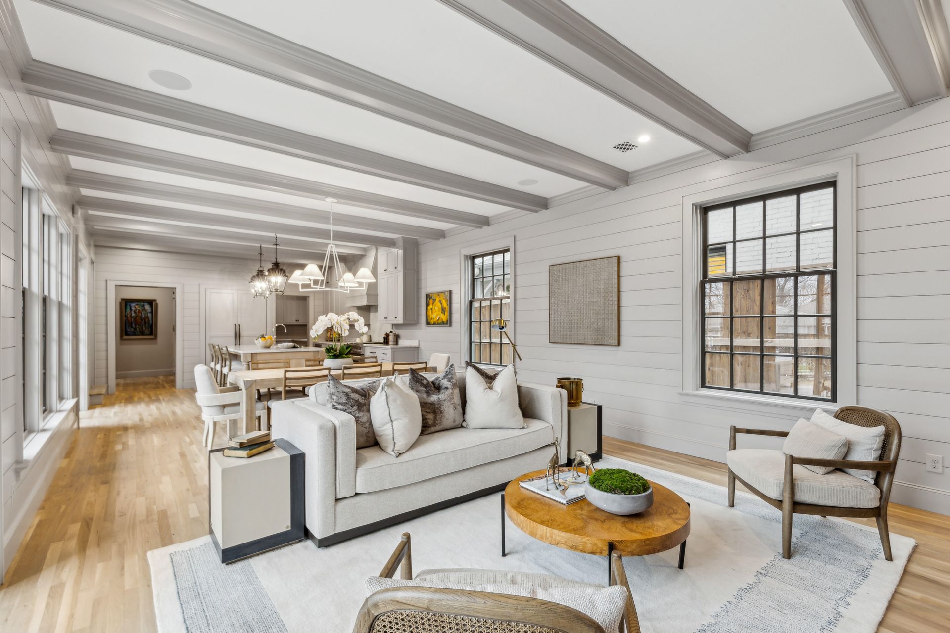 Living room with light-colored walls, exposed beams, open to a kitchen. Furnished with a sofa, chairs, and a round coffee table.