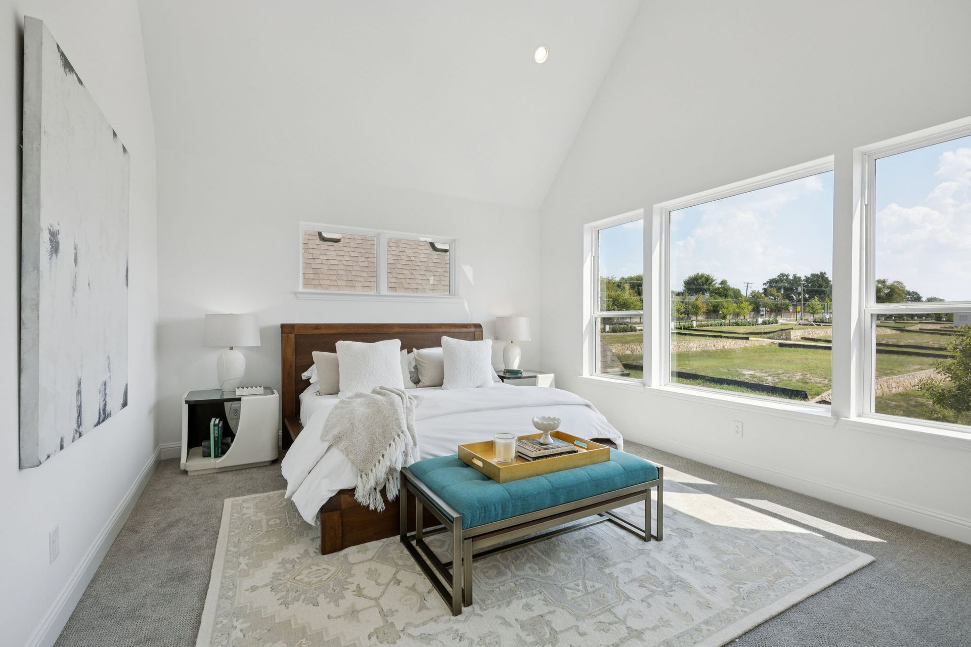 Bright bedroom with a bed, bench, large windows, and artwork. Soft colors and a view of green space.