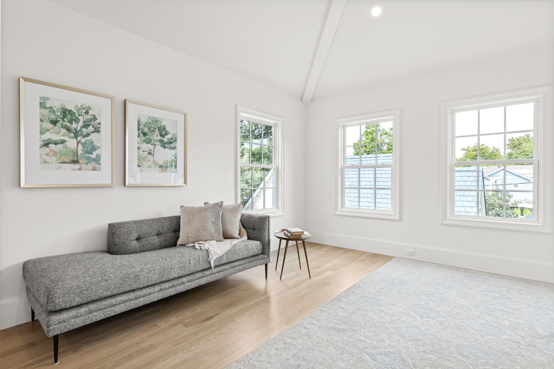 White room with gray chaise lounge, two framed art pieces, three windows, small table, and light wood floors.