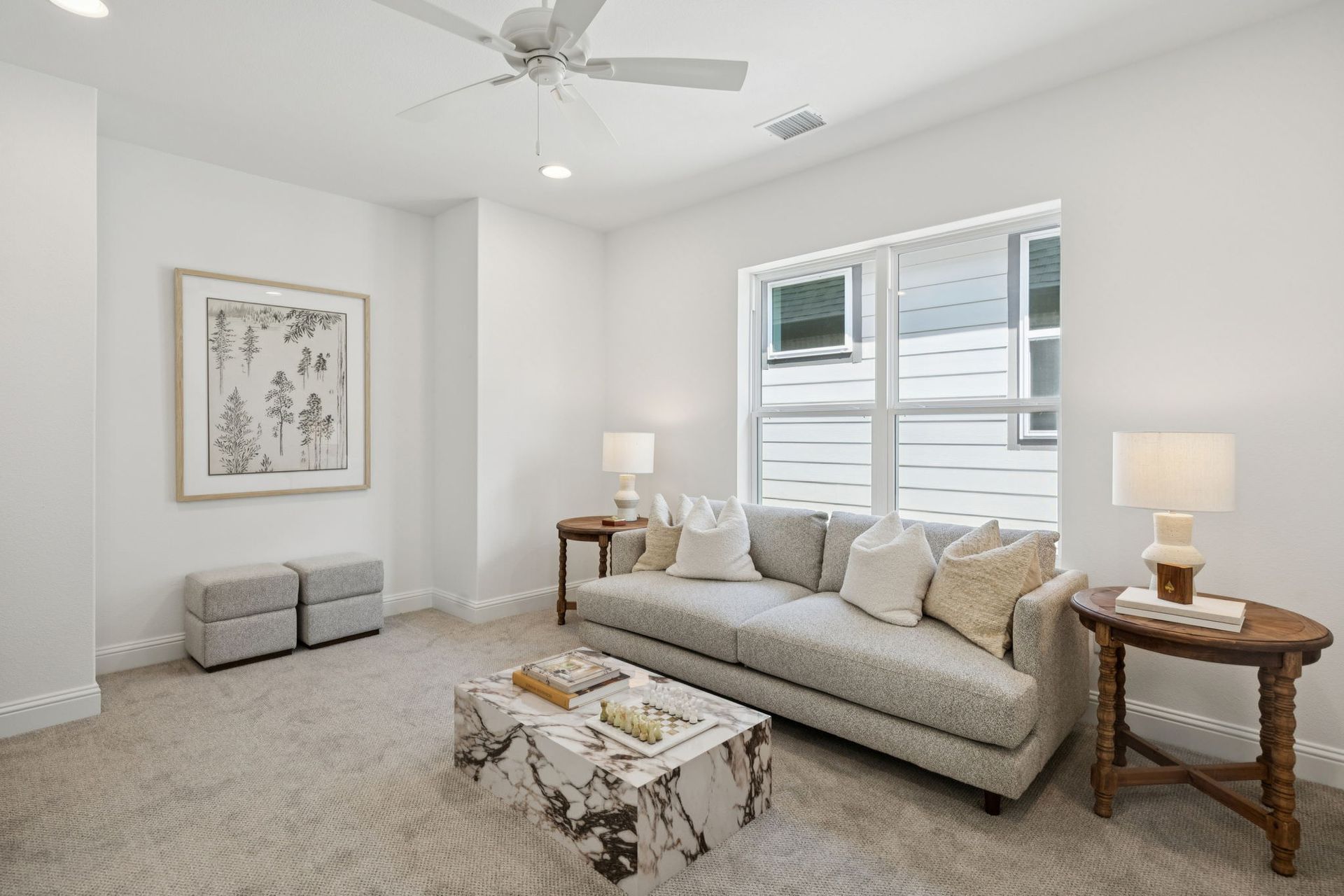 White-walled living room with gray sofa, small tables, and a marble coffee table. Window and framed art present.