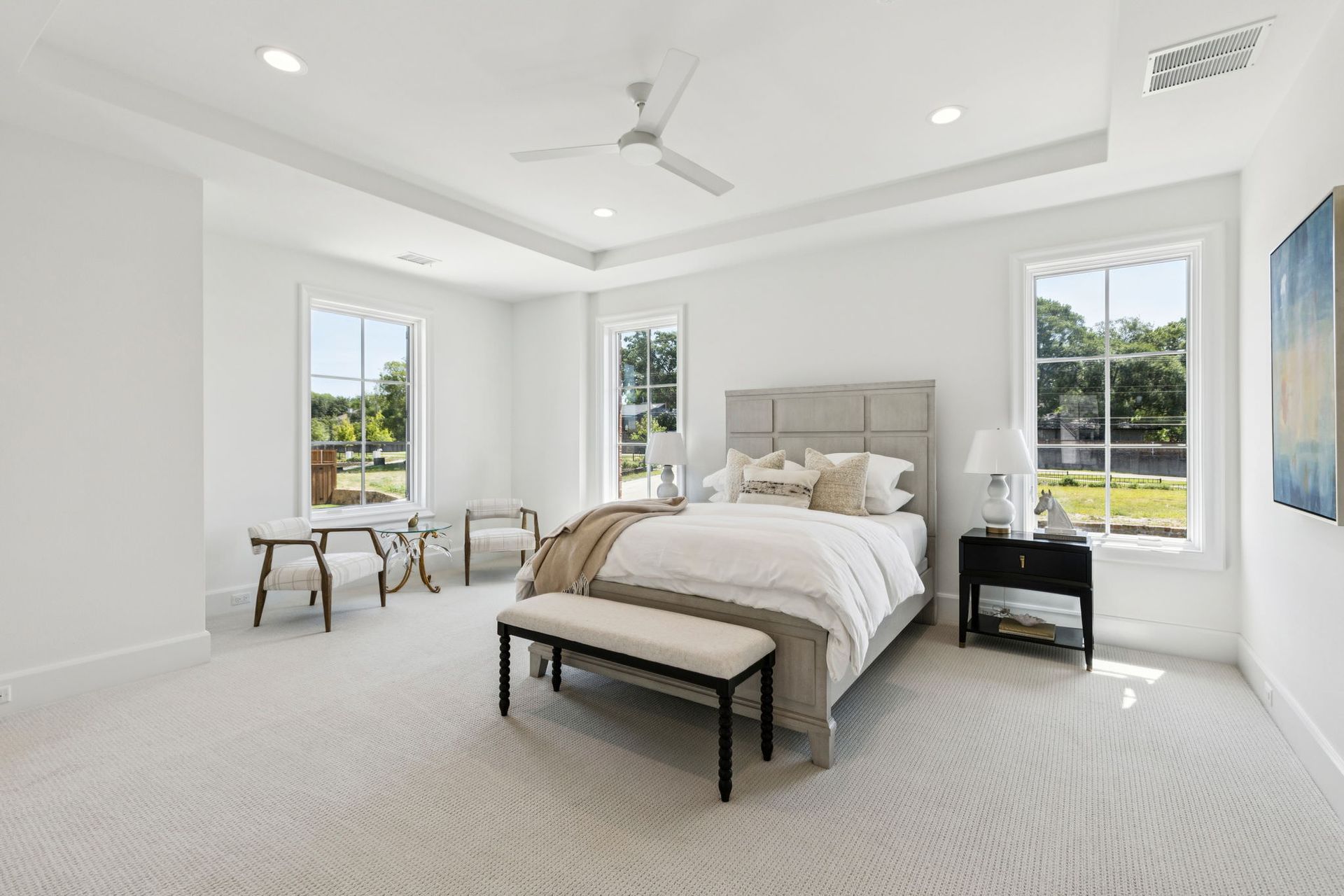 Bright, white bedroom with a bed, bench, chairs, and windows overlooking a green yard.