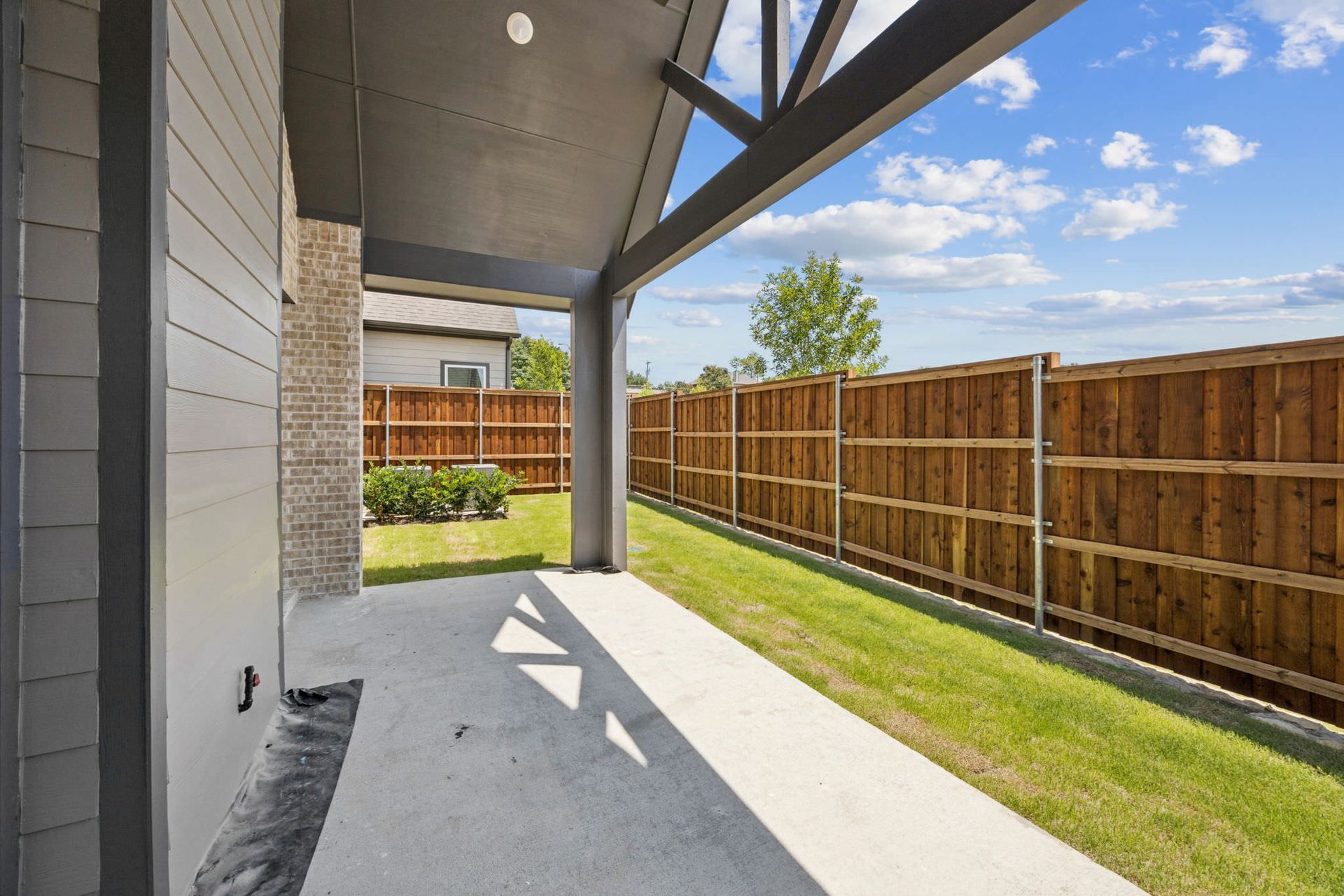 Covered patio with a concrete floor, green grass, and wooden fence on a sunny day.