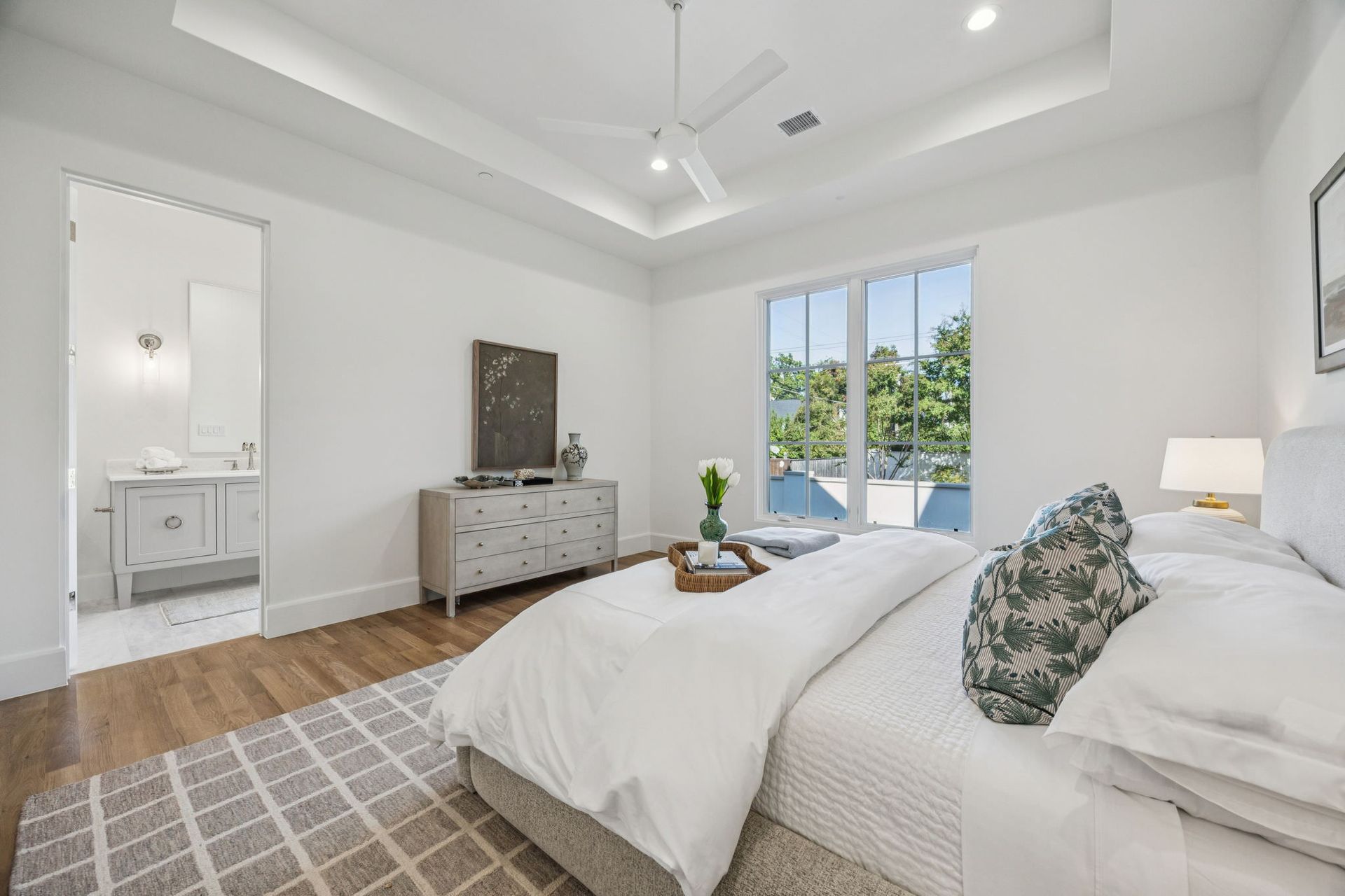 Bright bedroom with a bed, dresser, and a doorway to a bathroom. Large window with greenery.
