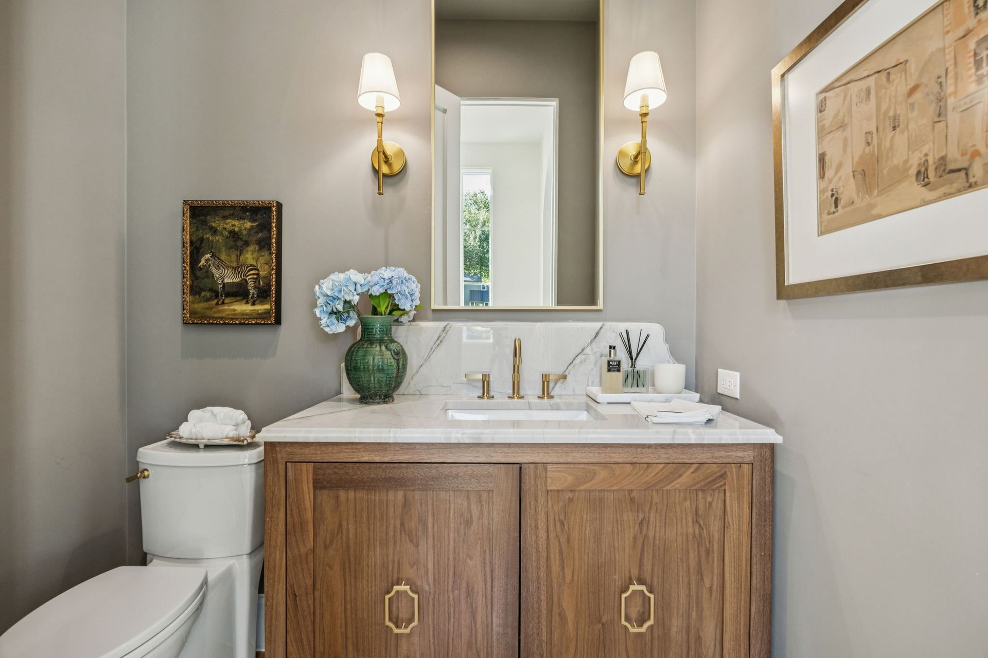 Powder room with wood vanity, marble countertop, gold accents, and artwork.