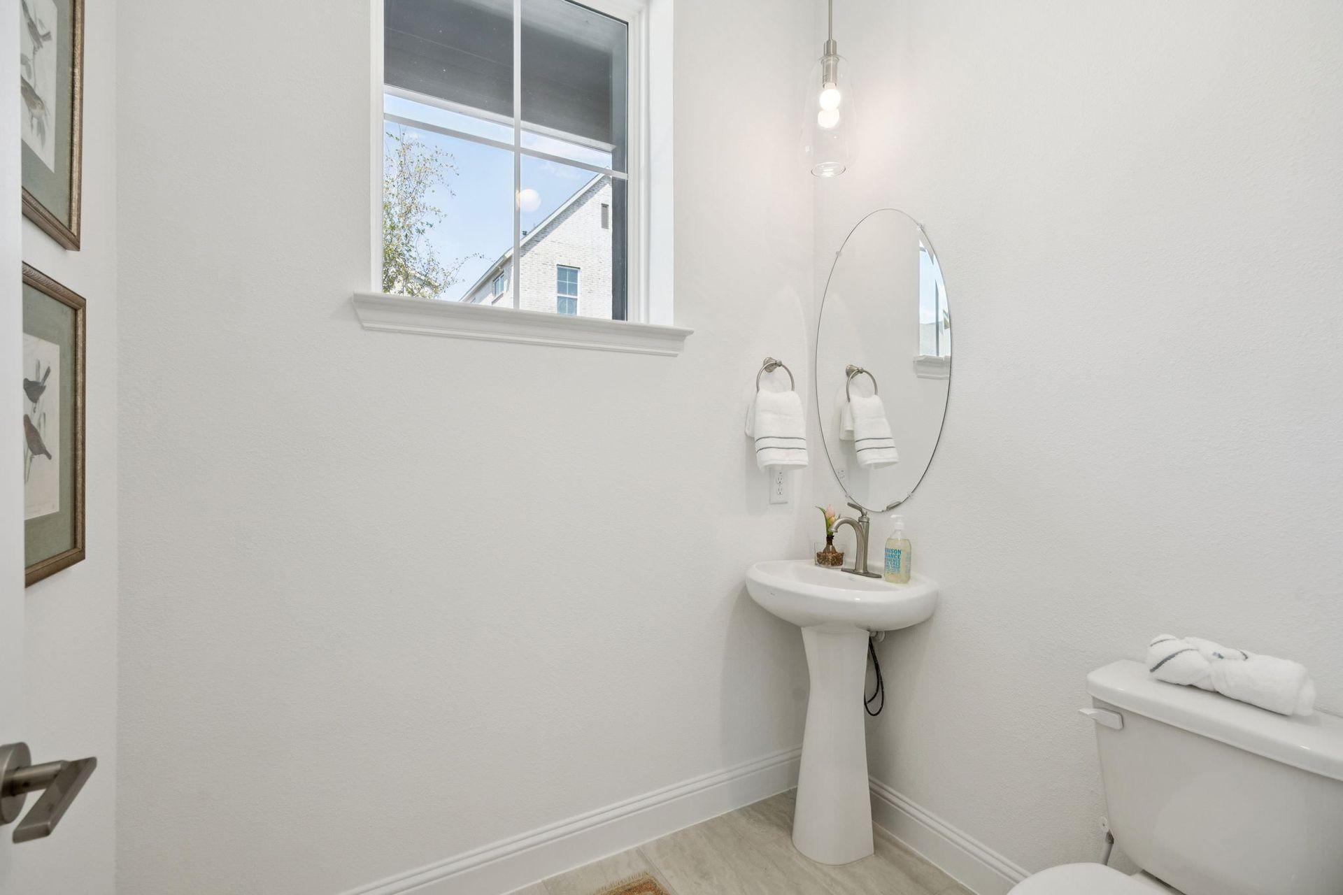 A small white bathroom with a pedestal sink, mirror, and toilet. A window is in the background.