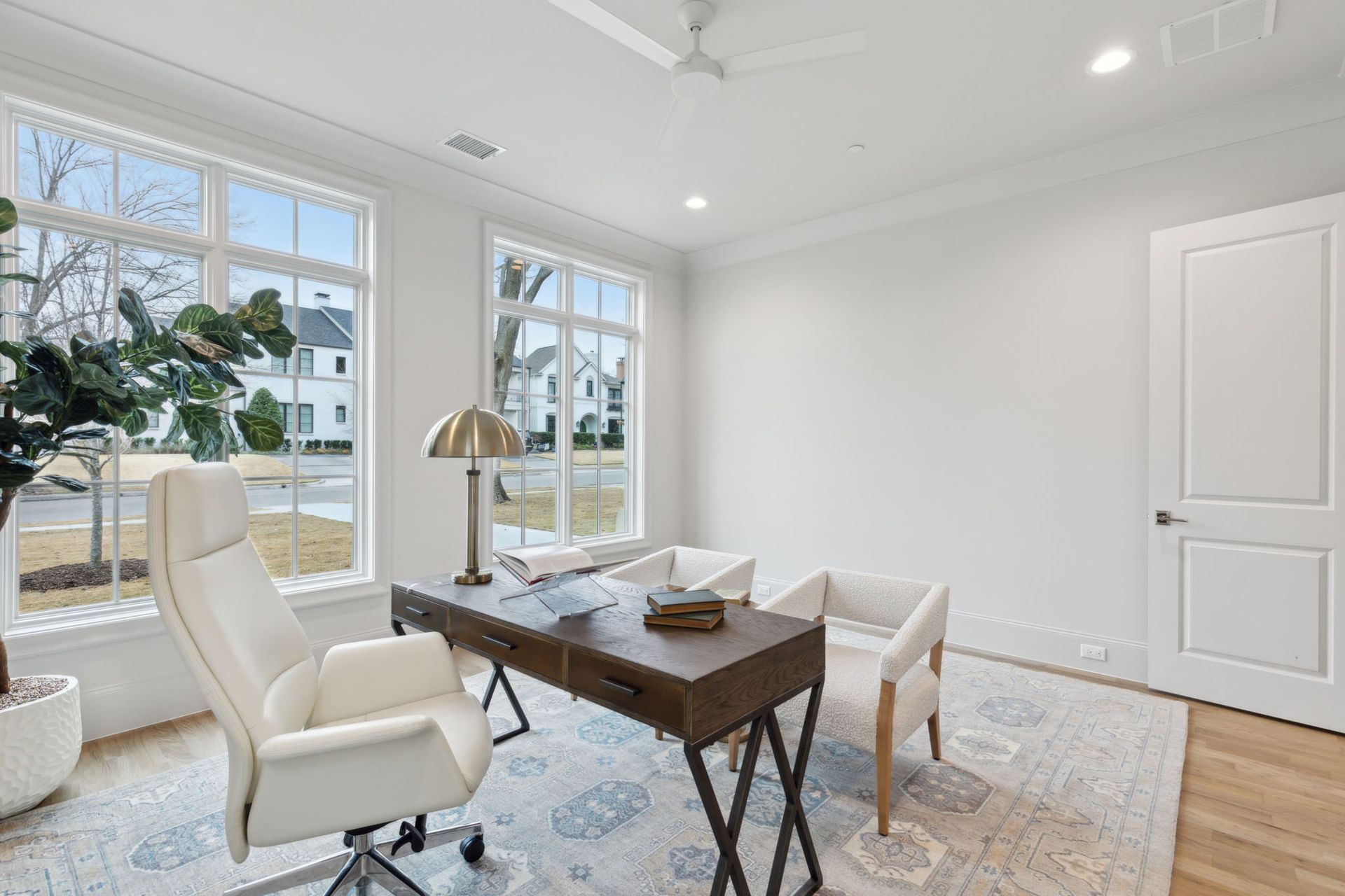 Home office with desk, two chairs, and large windows. White walls, gold lamp, light-colored rug, and a white door.