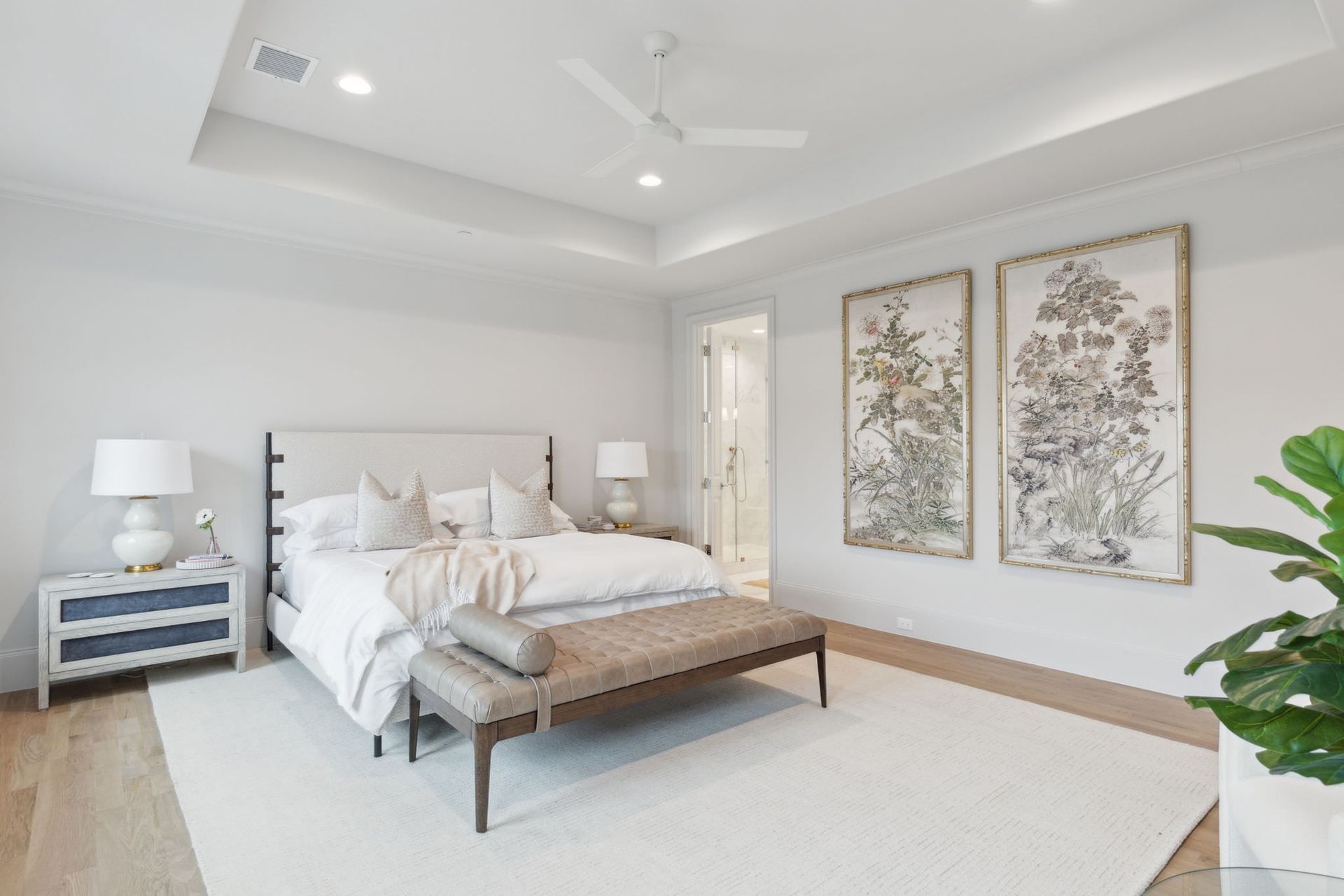 A bright bedroom with a bed, bench, artwork, and nightstands. Light wood floors and a white rug.