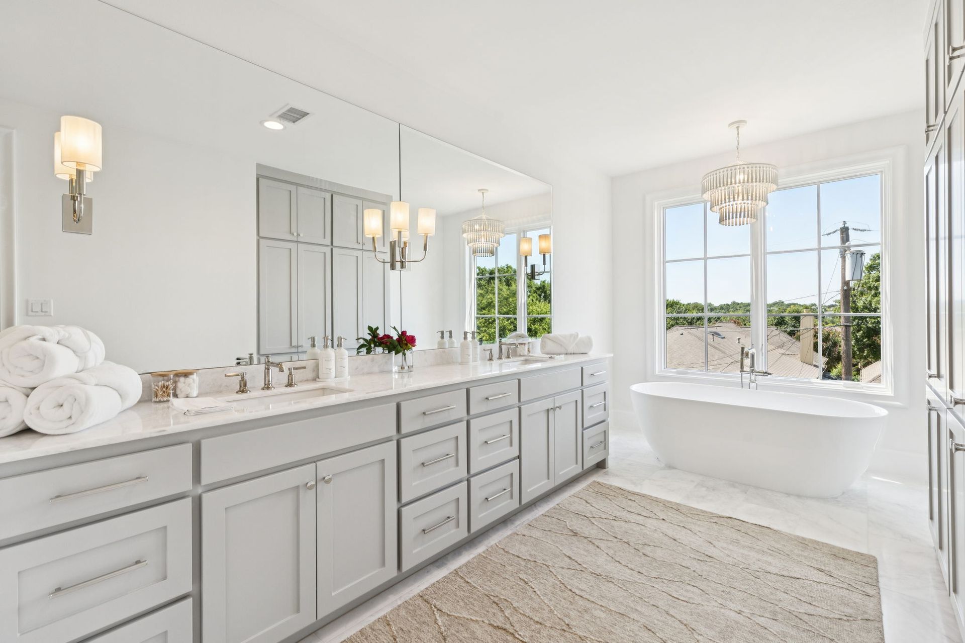 Spacious, bright white bathroom with gray cabinets, large mirror, and a freestanding tub by a window.