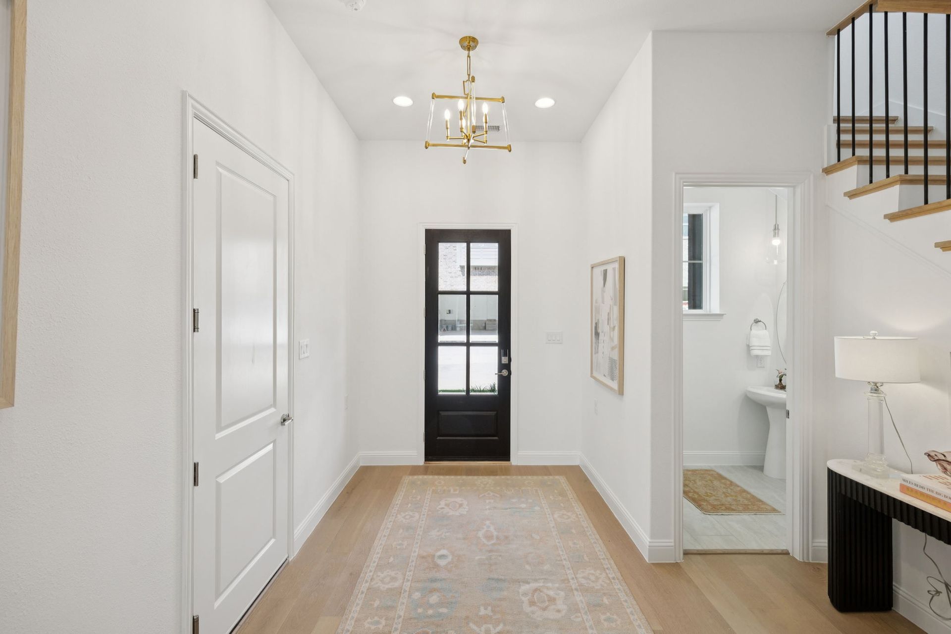 Hallway with white walls, wooden floor, black door, chandelier, and a rug.