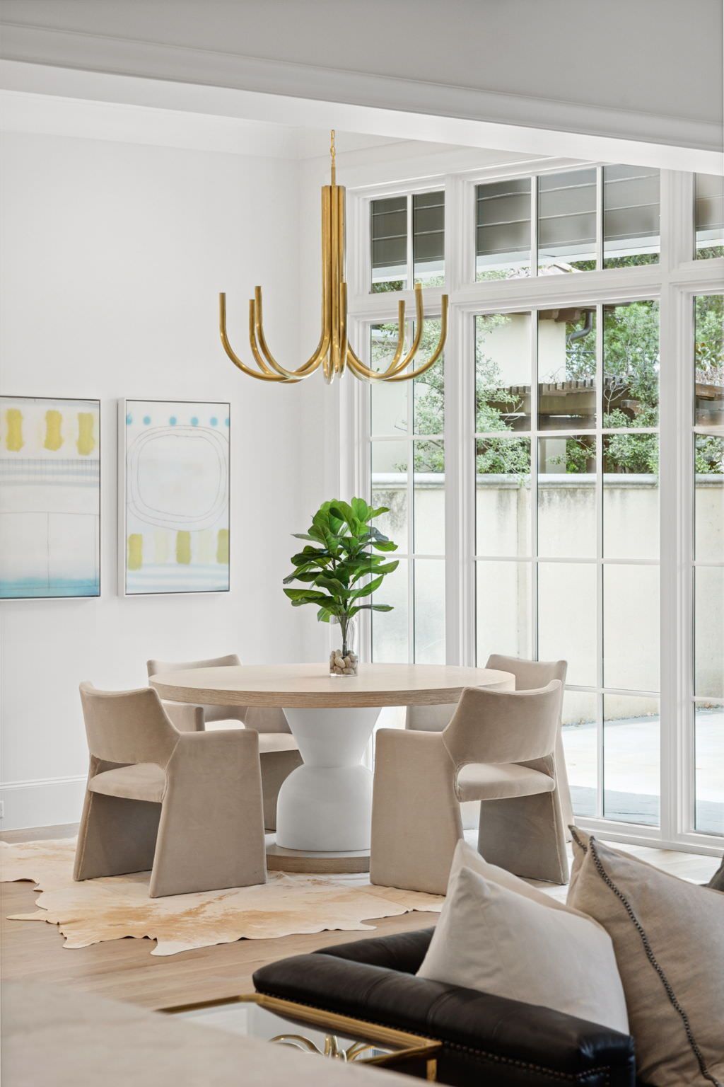 Dining area with round table, beige chairs, gold chandelier, and large windows.
