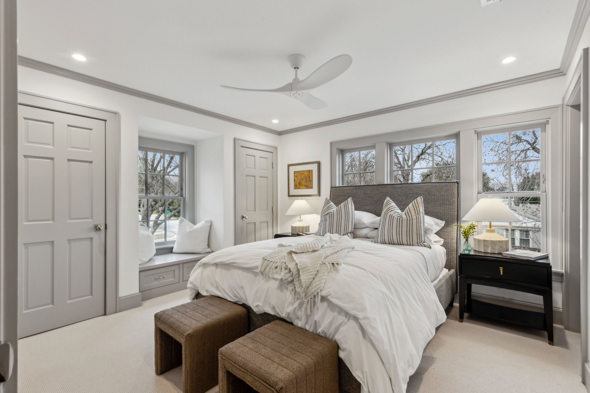 Bedroom with bed, window seat, neutral tones, gray trim, and two woven stools.