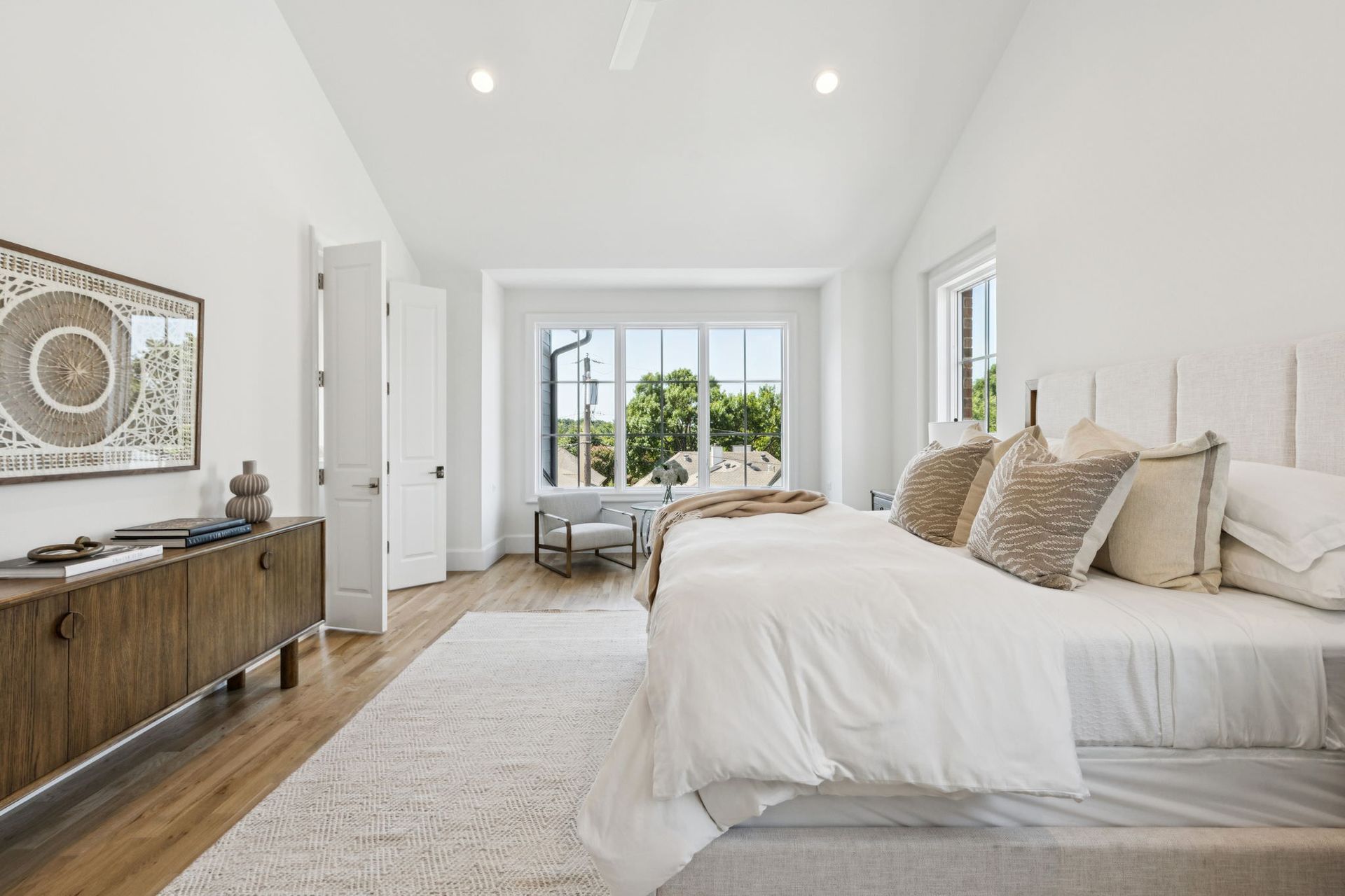 Bright bedroom with a large bed, rug, wooden dresser, and large window with outside view.
