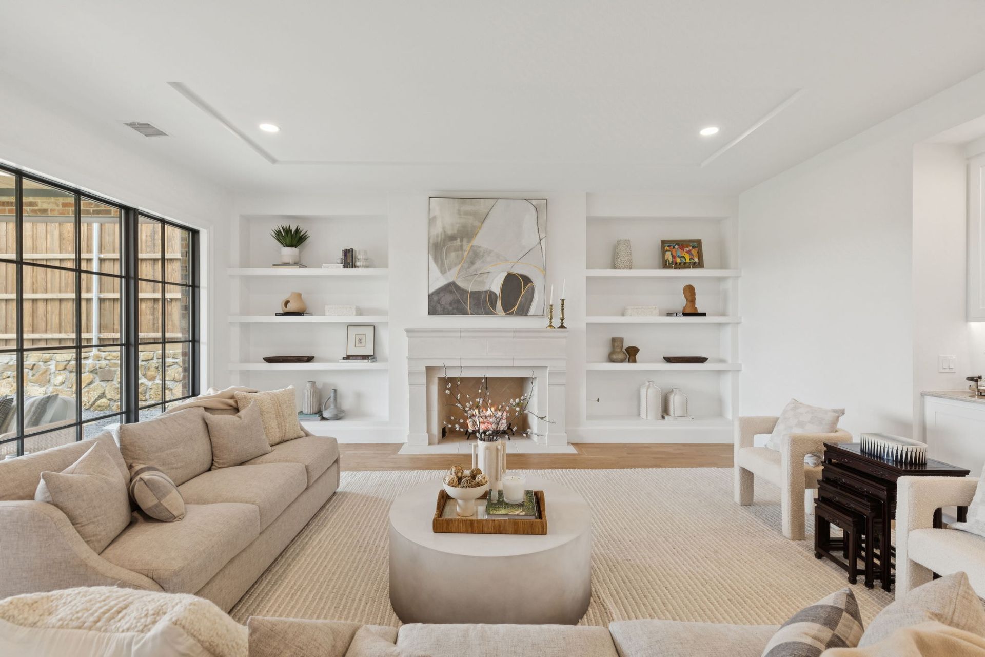 Bright living room with built-in bookshelves flanking a fireplace. Beige sofa, round coffee table, and large window.